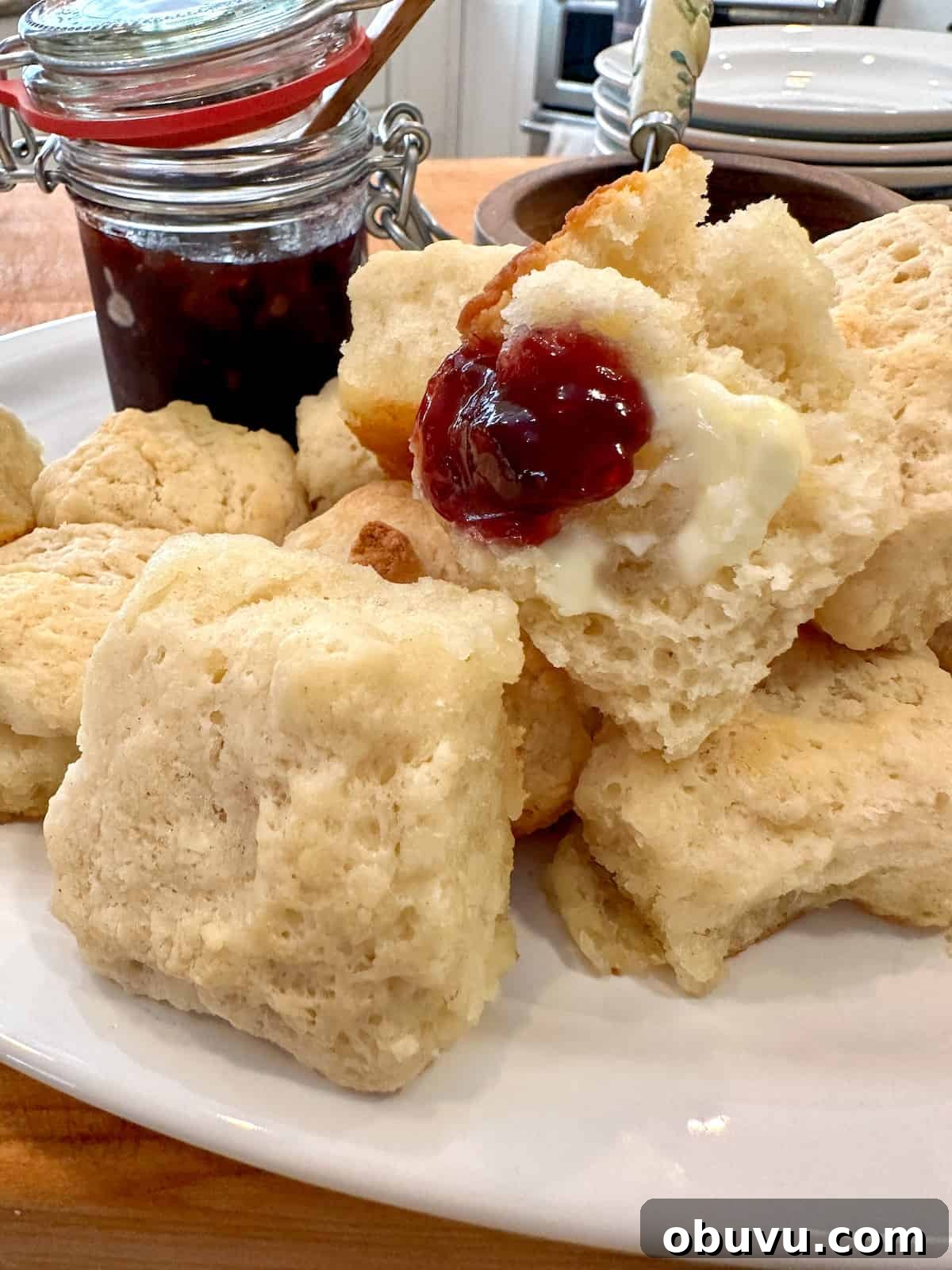 Flaky Buttermilk Biscuits Recipe for Perfect Homemade Biscuits 2 Biscuits on a plate with a portion of jam on one biscuit and a jar of jam.