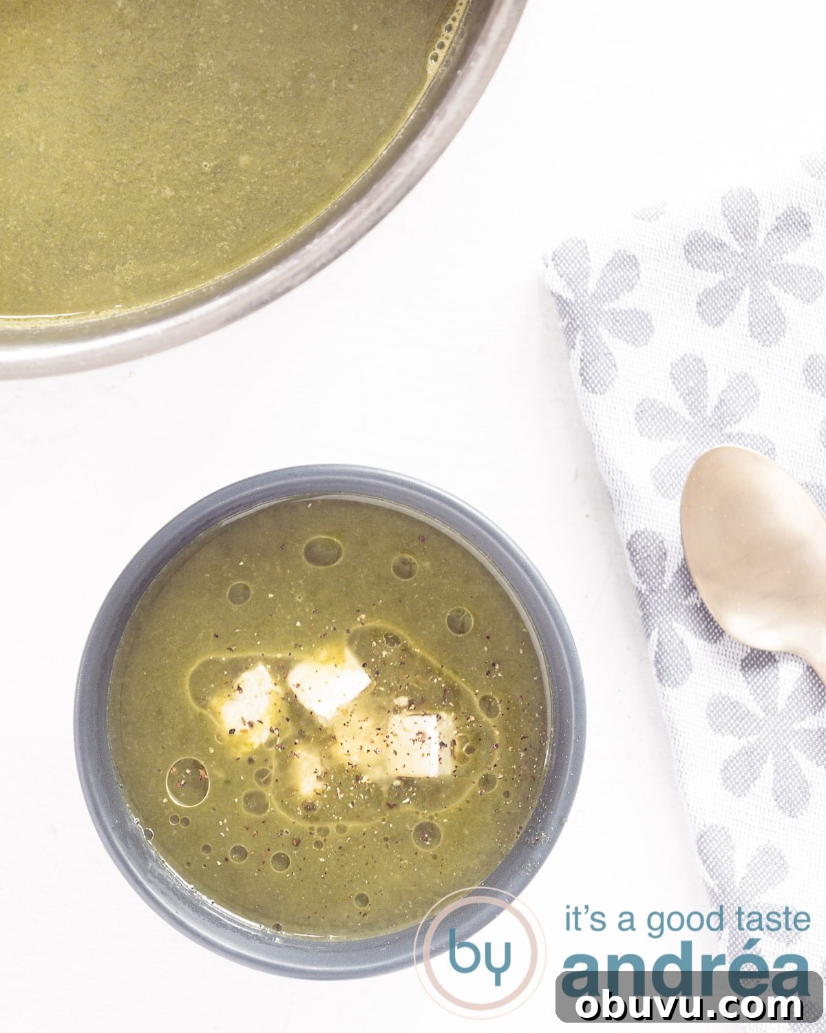 Creamy Vegan Spinach and Potato Soup Recipe 2 A photo from above showing a bowl of soup with spinach, potato and onion. On the right side a gray and white tea towel, a golden spoon. Part of a pan of soup at the top. Everything on a white background