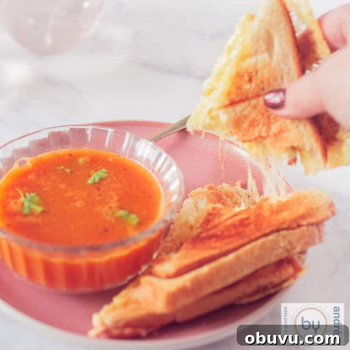 A square photo of a bowl of tomato soup with parsley and a grilled cheese sandwich with a piece broken off, revealing streaks of melted cheese