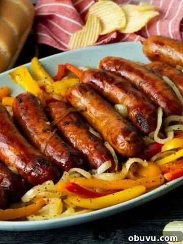 Grilled sausage atop onions and peppers on blue platter with buns and potato chips in background.