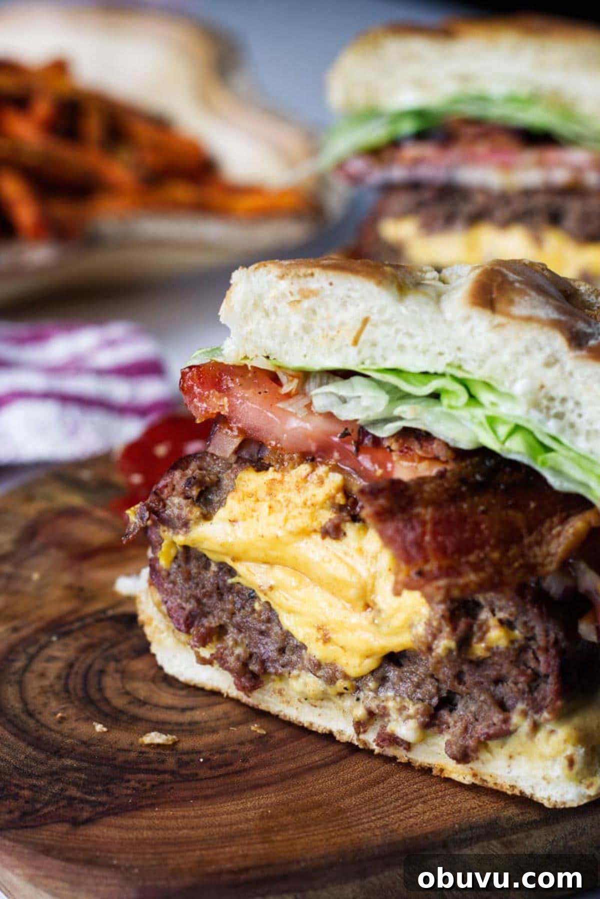 Juicy Lucy Burger Recipe: Melted Cheese-Stuffed Patties 2 A Juicy Lucy burger cross section, sitting on a wood cutting board, cheese spilling out the middle with bacon, onion, tomato, lettuce, with sweet potato fries in background