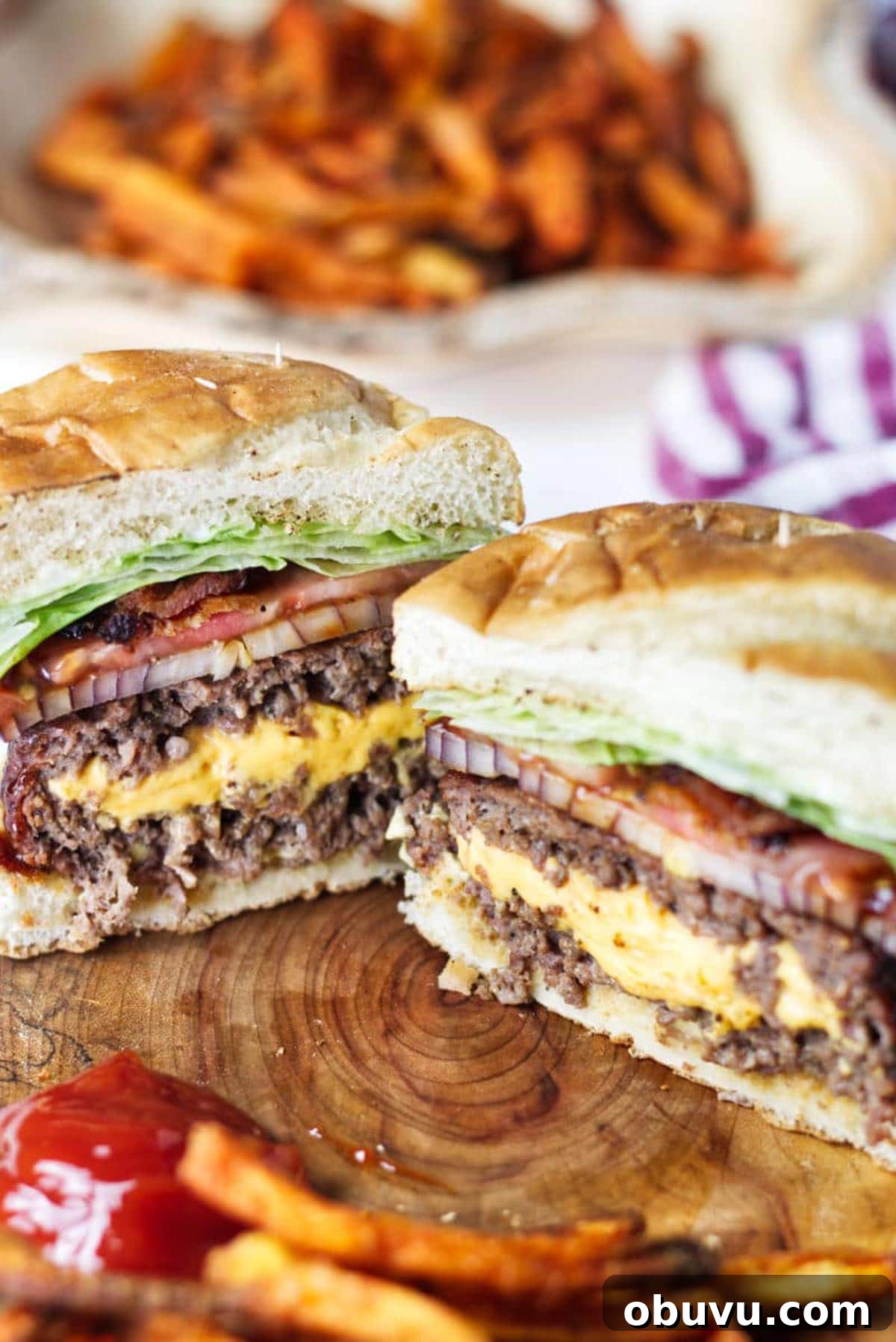 Juicy Lucy Burger Recipe: Melted Cheese-Stuffed Patties 3 A Juicy Lucy burger cross section, sitting on a wood cutting board, cheese spilling out the middle with bacon, onion, tomato, lettuce, sweet potato fries and ketchup