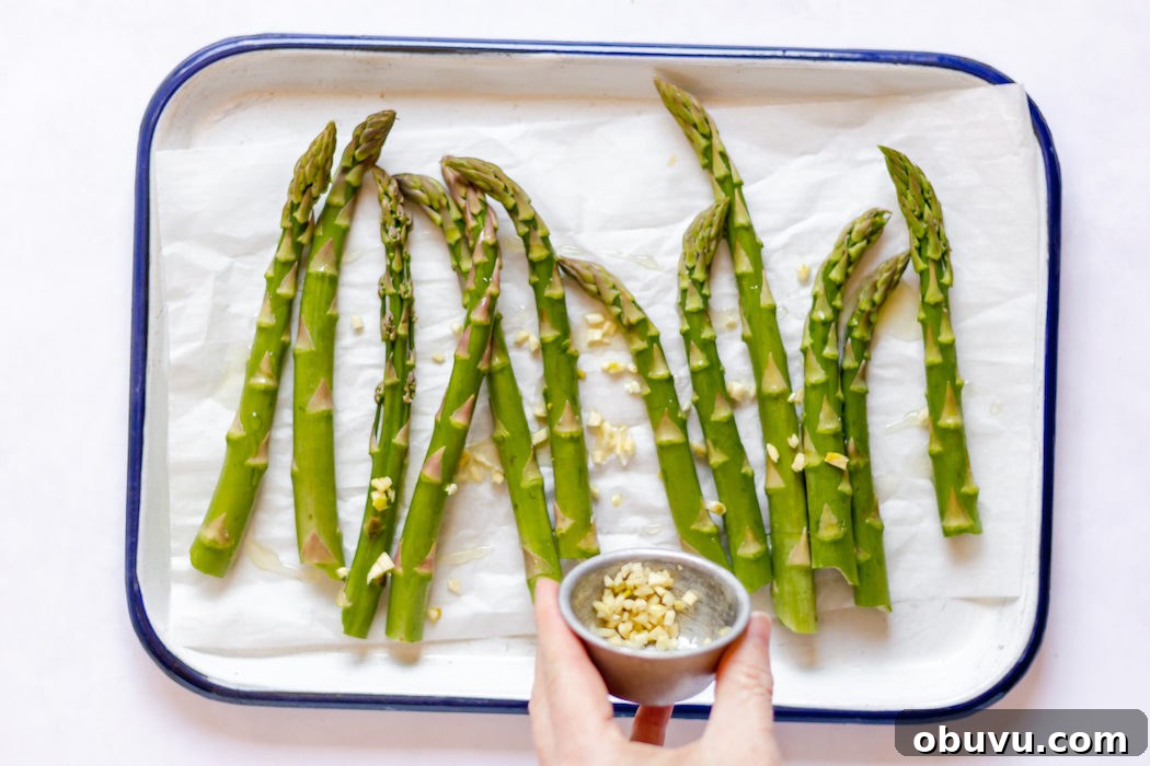 Image, taken from above, shows a tray of asparagus and a hand sprinkling diced garlic on the stalks.