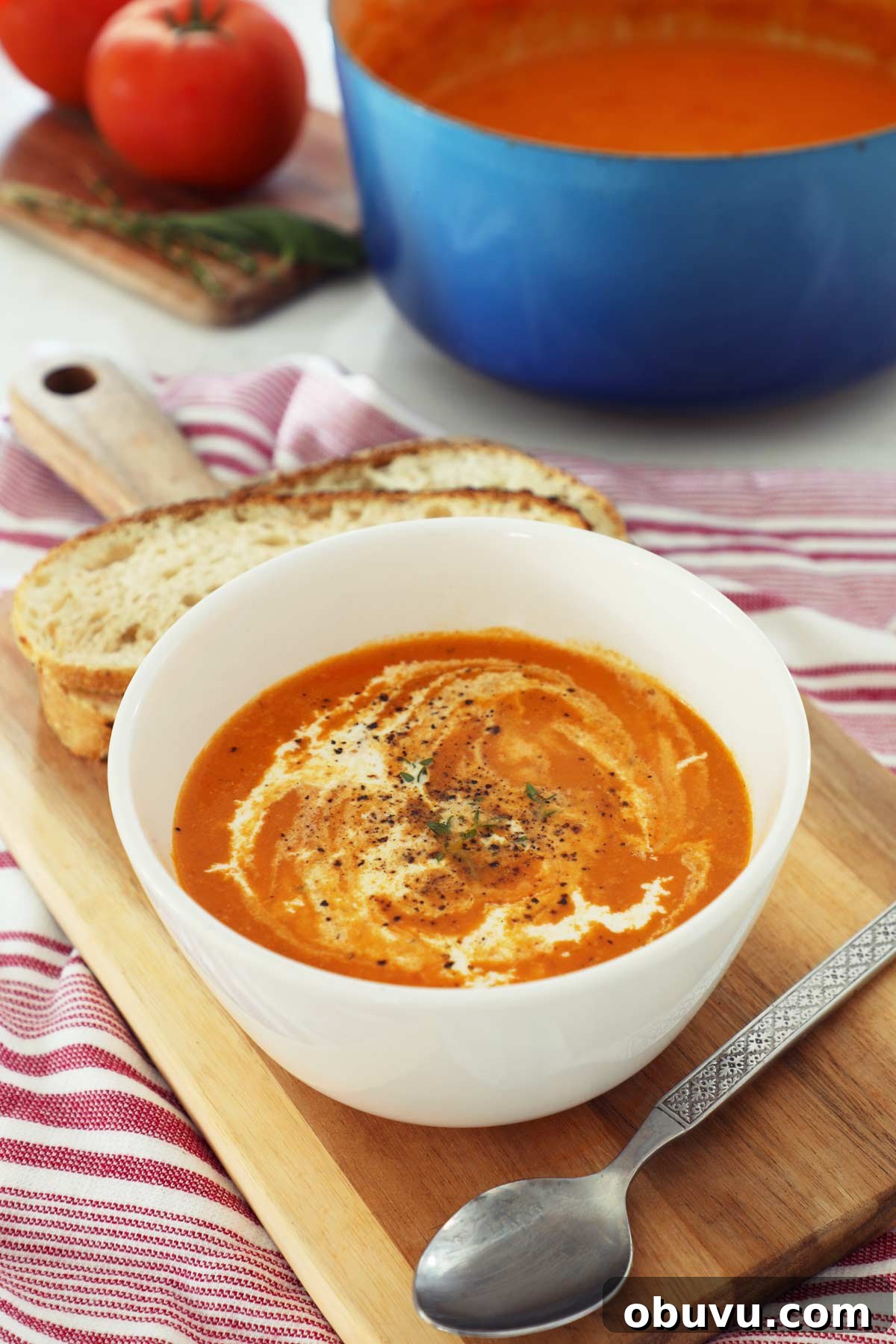 Roasted Red Pepper and Tomato Soup Recipe — Cozy Weeknight Dinner 2 A white bowl of roasted tomato and red pepper soup topped with cream on a cutting board with slices of bread and a dutch oven of soup in the background.