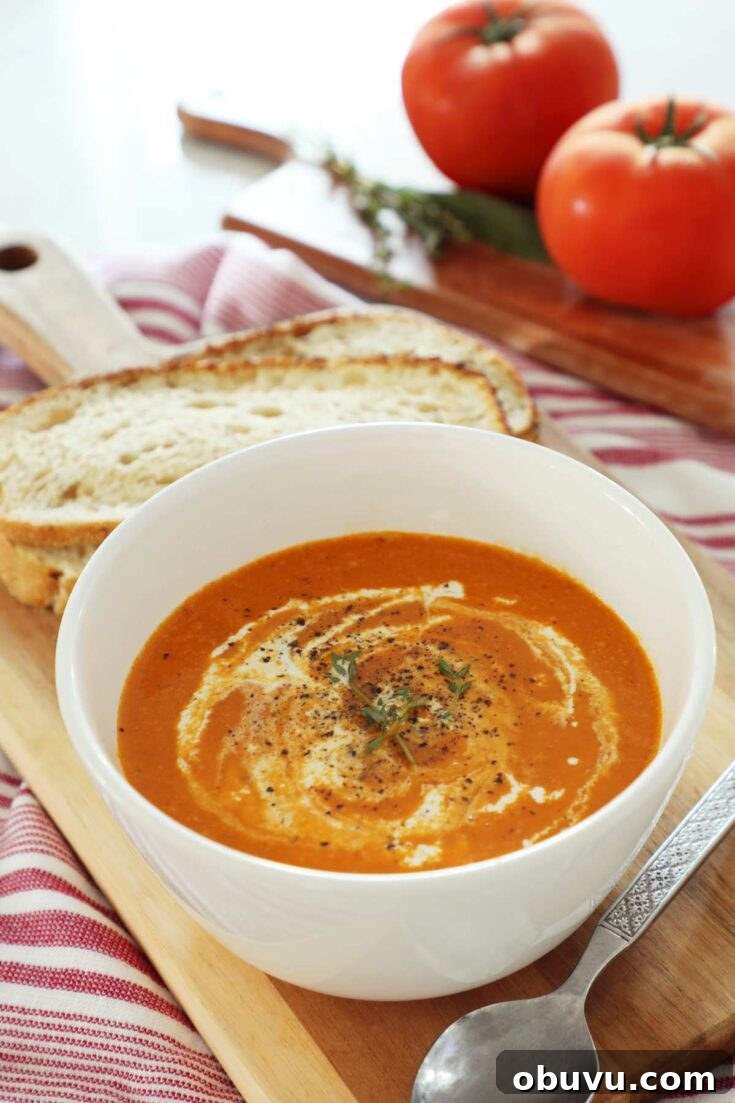 Roasted tomato and red pepper soup in a white bowl with a creamy swirl on top, along with some bread sits on a wooden board on top of the kitchen counter, with tomatoes in the background.