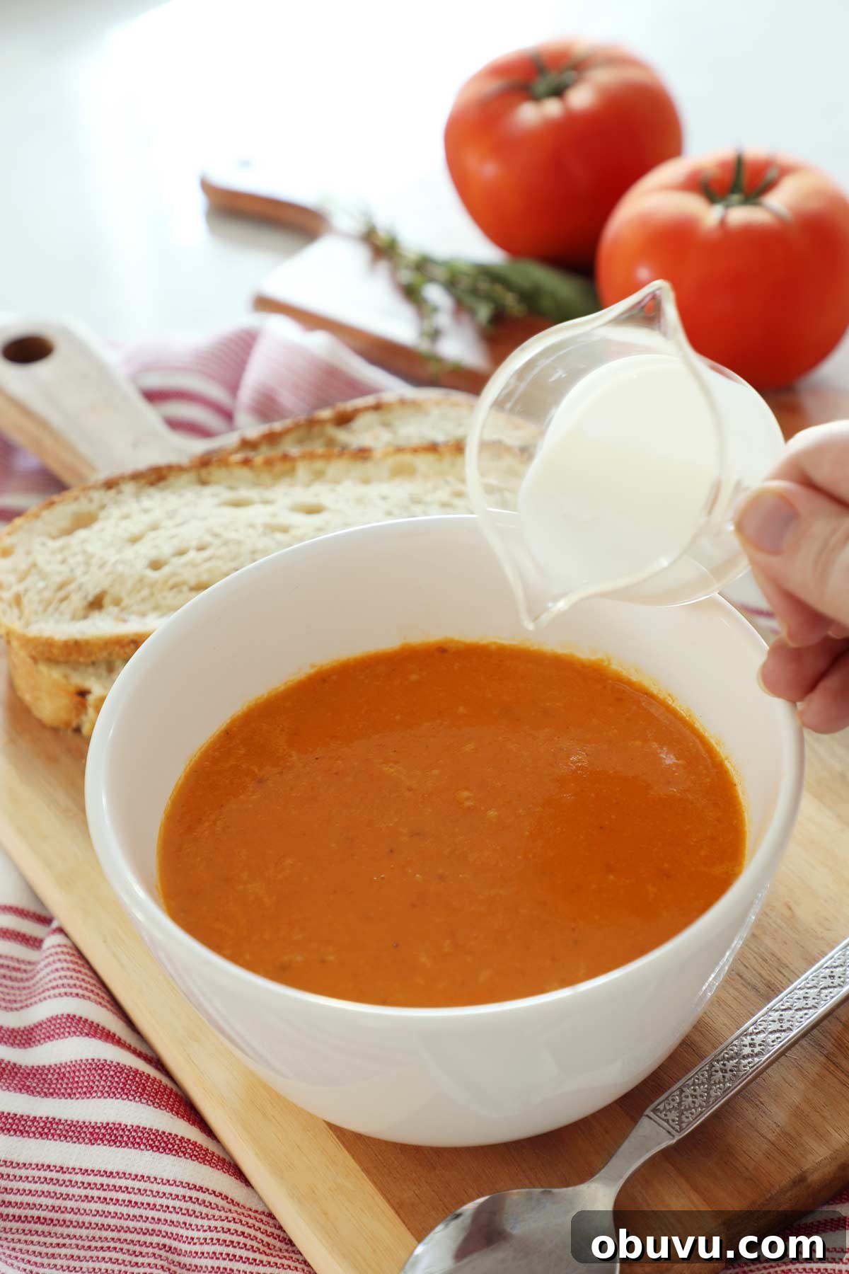 Roasted Red Pepper and Tomato Soup Recipe — Cozy Weeknight Dinner 7 A white bowl filled with tomato and roasted red pepper soup with cream being poured in.