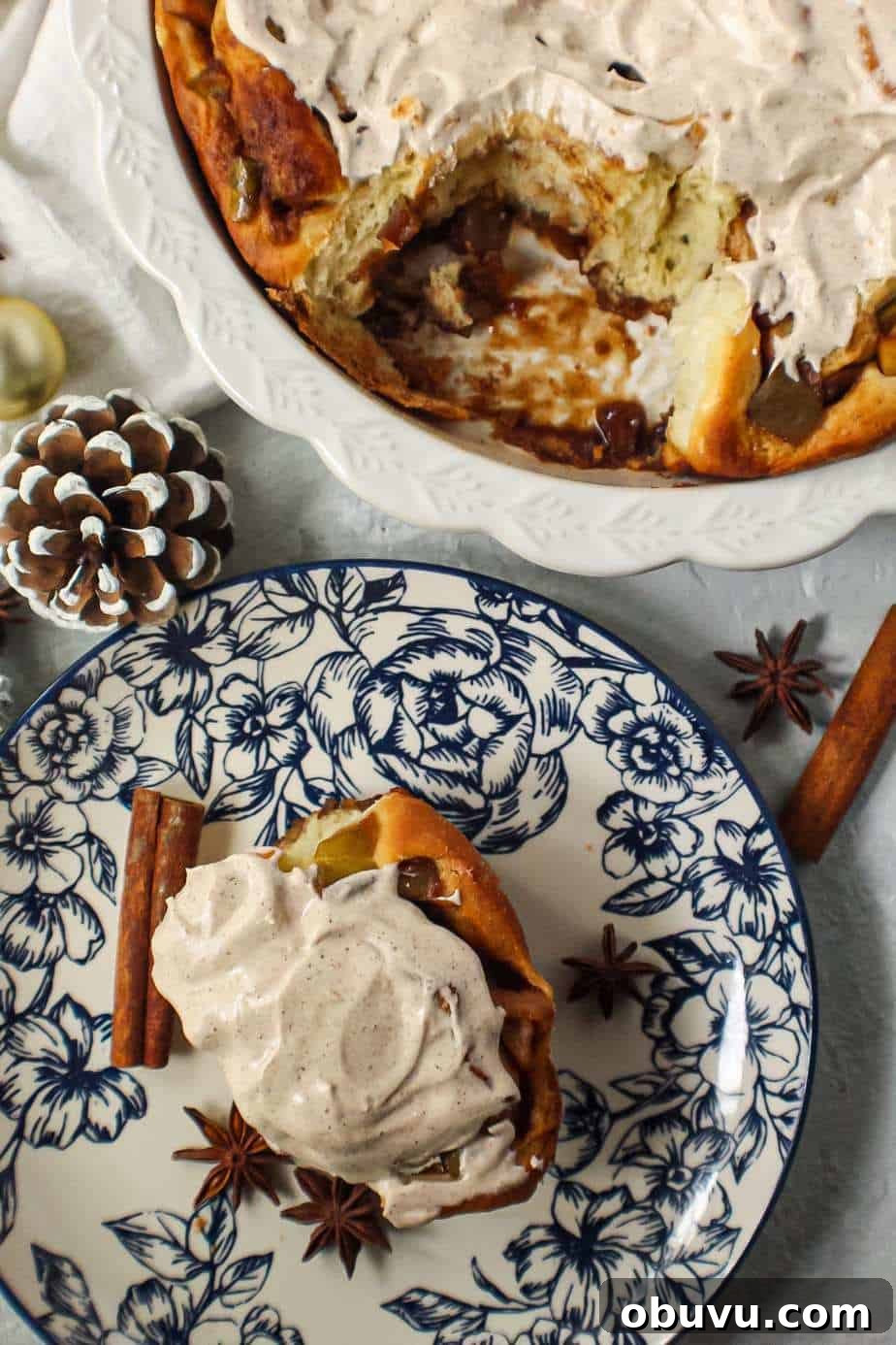 Apple Cinnamon Rolls with Chai-Spiced Cream Cheese Glaze 3 apple cinnamon rolls with chai cream cheese frosting on top in white pie dish with one cinnamon roll on a blue and white plate next to pie dish with cinnamon sticks and star anise in shot.