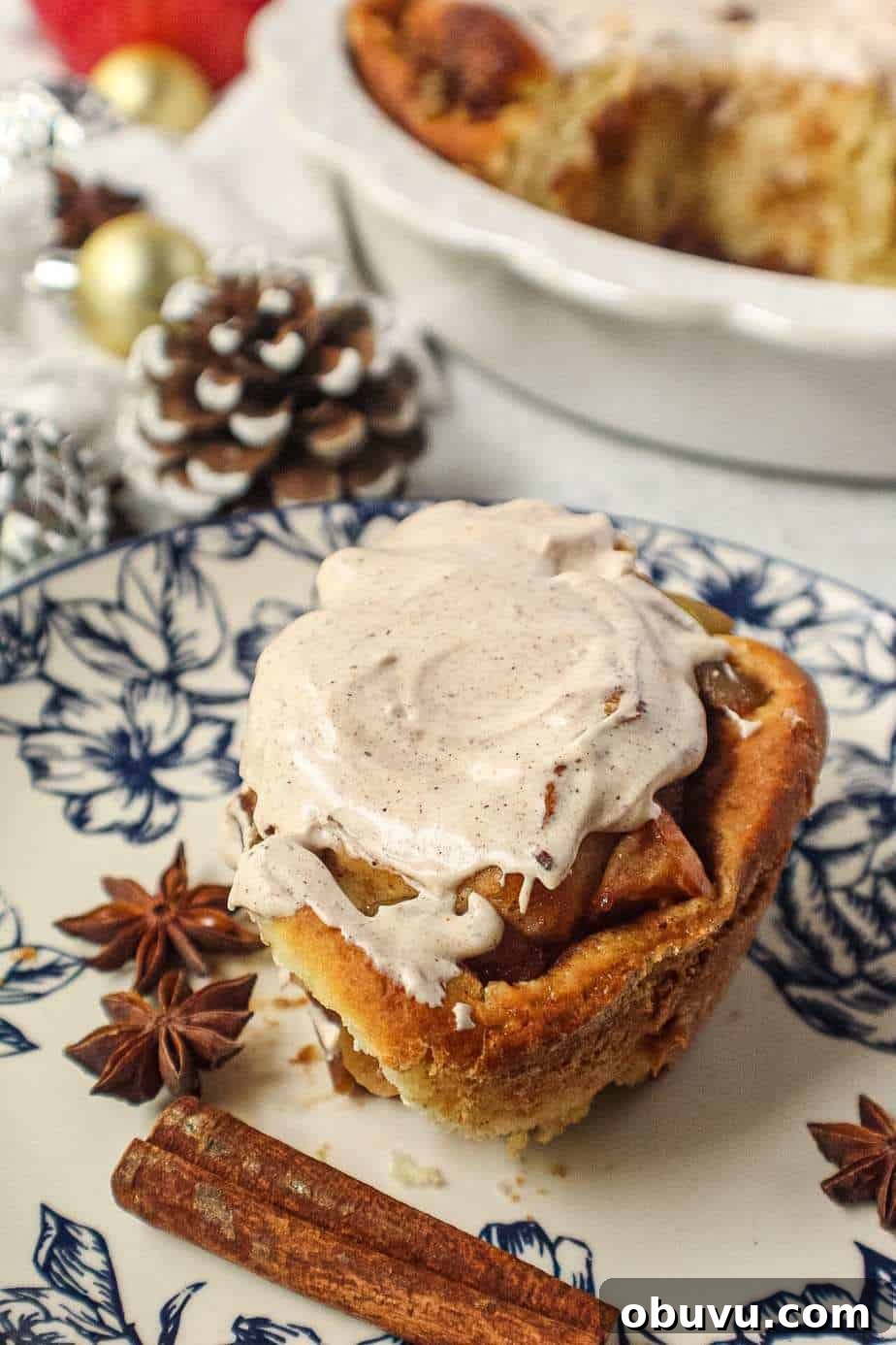 Apple Cinnamon Rolls with Chai-Spiced Cream Cheese Glaze 6 apple cinnamon rolls with chai cream cheese frosting on top in white pie dish with one cinnamon roll on a blue and white plate next to pie dish with cinnamon sticks and star anise in shot.
