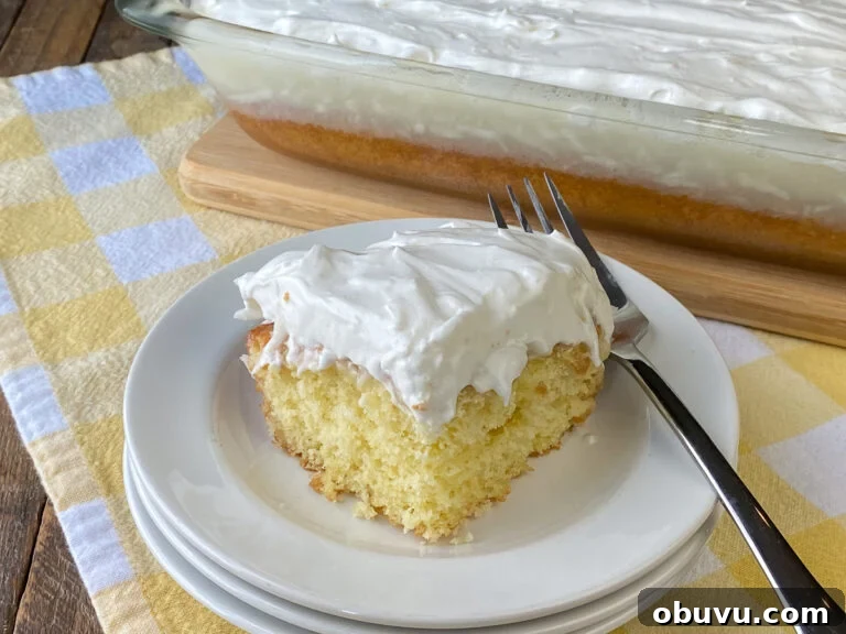 Bright piece of sour cream coconut cake with thick icing on a white plate with a fork in front of the 9x13 pan full of coconut cake