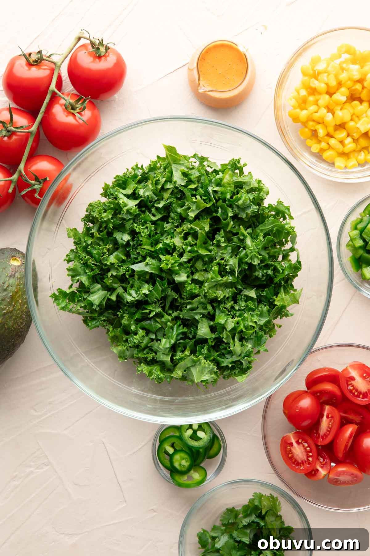 Chipotle Kale Salad Recipe with Smoky Lime Dressing 4 bowl of massaged kale surrounded by small glass bowls of salad ingredients: corn, jalapenos, bell peppers, tomatoes, cilantro, and chipotle dressing