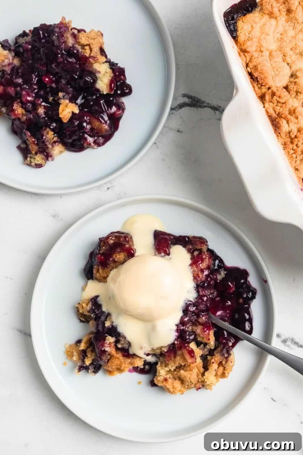 Blueberry Cobbler Dump Cake Made with Muffin Mix 3 overhead view of two white plates with helpings of blueberry cobbler dump cake, one of which is topped with a scoop of vanilla ice cream.