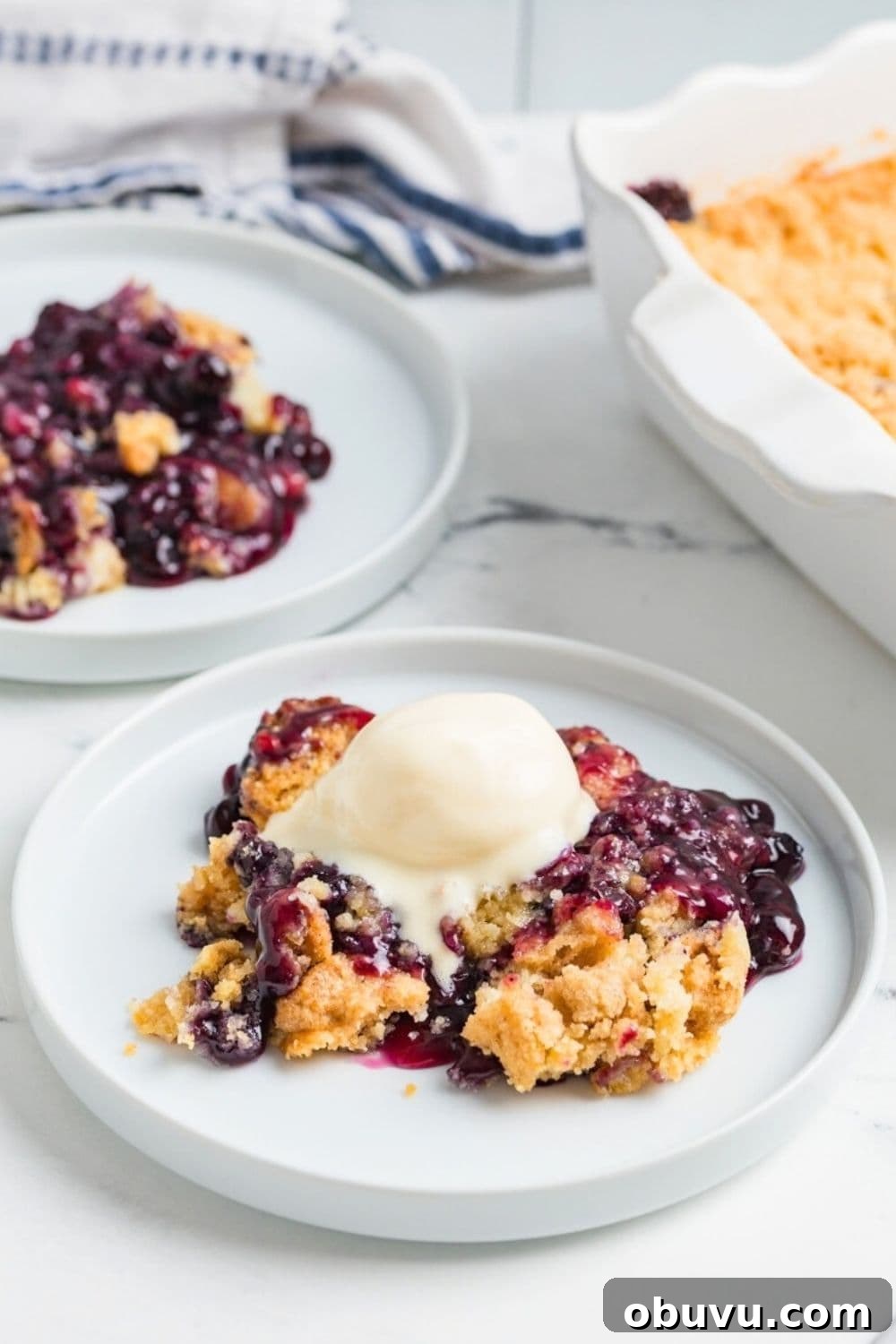 Blueberry Cobbler Dump Cake Made with Muffin Mix 9 helpings of blueberry cobbler made with muffin mix served on white plates. A scoop of ice cream melts over one of the servings of warm cobbler.