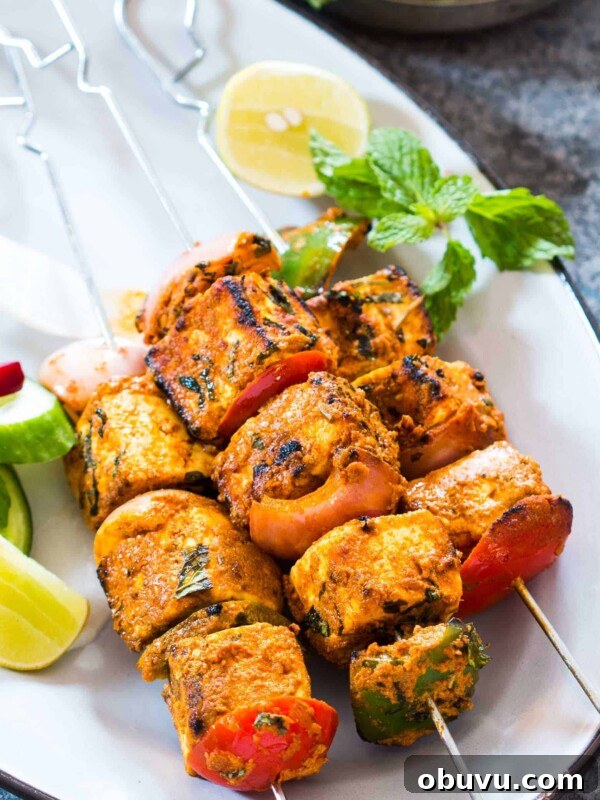Paneer tikka skewers garnished with mint, lemon and served on a white plate.
