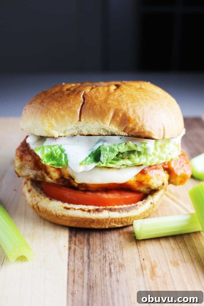 Spicy Buffalo Chicken Breast Burger Recipe 2 A Buffalo chicken burger topped with ranch dressing, tomato and lettuce on a wooden cutting board