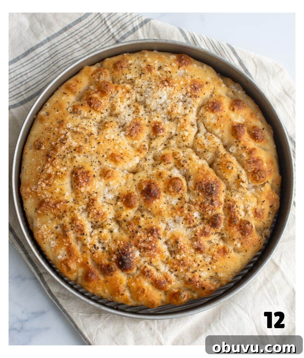 Sourdough Focaccia: Crispy, Airy Italian-Style Loaf Recipe 10 A round metal pan with golden brown baked sourdough focaccia topped with herbs and flaky sea salt.
