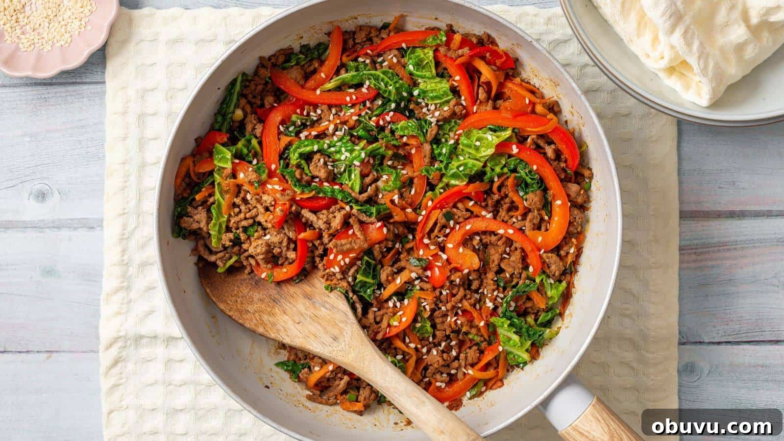 29 Delicious Low-Carb Meals That Aren’t Just Salad 29 A skillet containing a stir-fried mix of ground meat, sliced bell peppers, leafy greens, and white sesame seeds, with a wooden spoon resting on the side. Plates with flatbreads are in the background.
