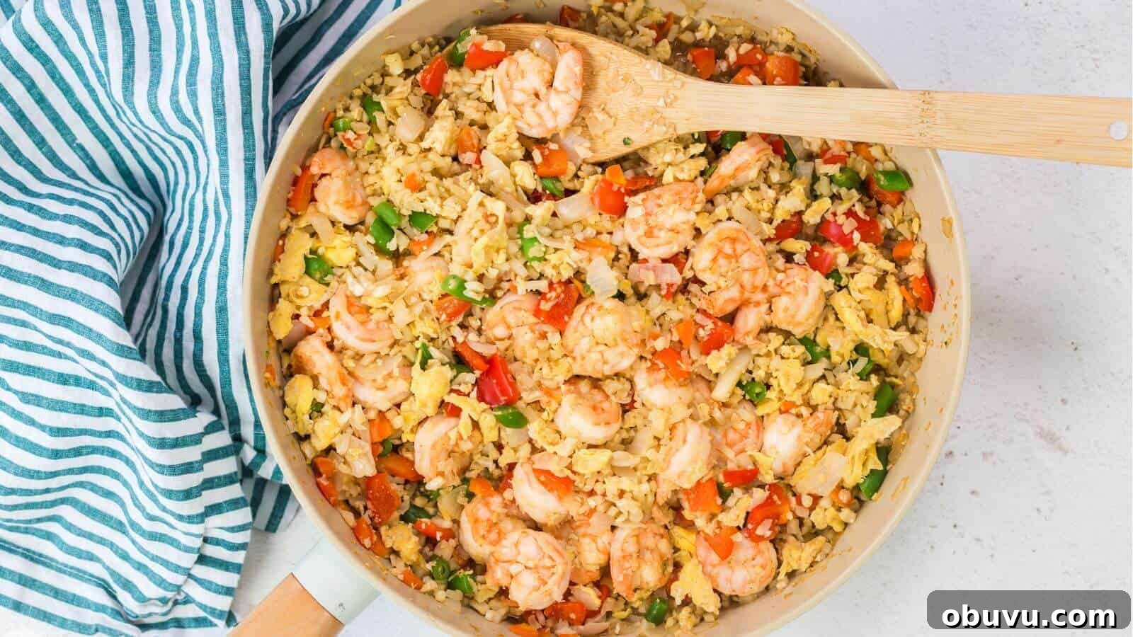 29 Delicious Low-Carb Meals That Aren’t Just Salad 5 A skillet brimming with keto fried cauliflower rice, shrimp, peas, carrots, and bell peppers is accompanied by scrambled eggs. A wooden spoon rests on the side, while a striped cloth adds charm next to the skillet.