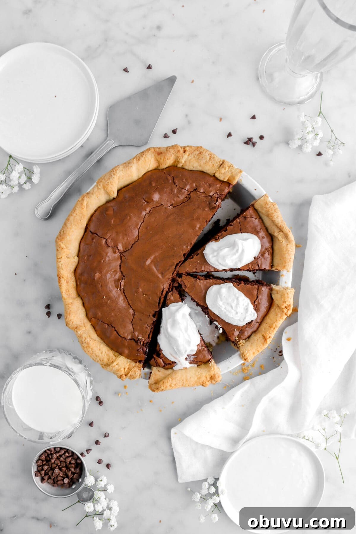 Fudgy Chocolate Chip Brownie Pie Recipe for Ooey-Gooey Dessert 2 overhead shot of brownie pie with three slices cut into pie with all three slices having a dollop of whipped cream on top, a white napkin and flowers around, with chocolate chips and a glass of milk beside on marble surface.