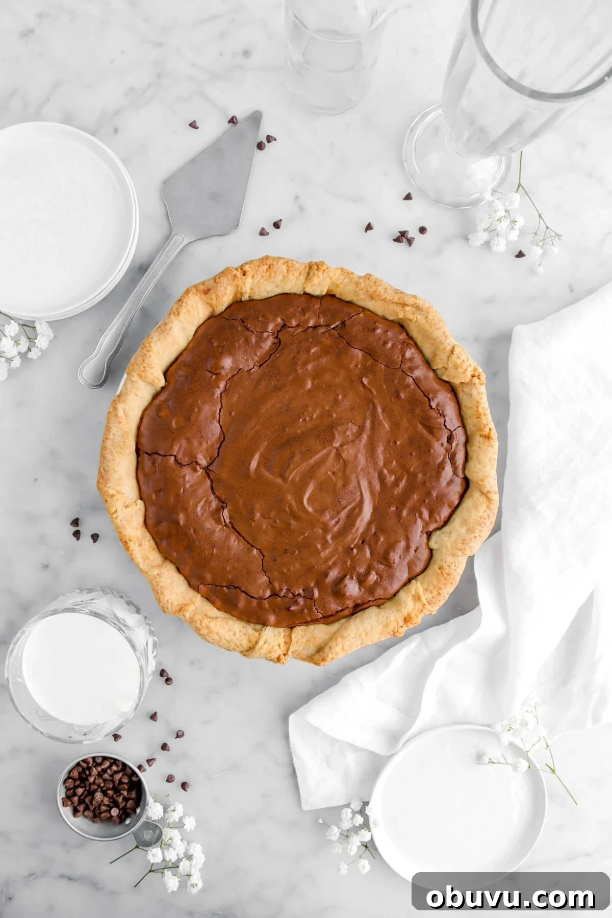 Fudgy Chocolate Chip Brownie Pie Recipe for Ooey-Gooey Dessert 4 brownie pie on marble surface with chocolate chips, white flowers, a white napkin, and a cake knife beside.