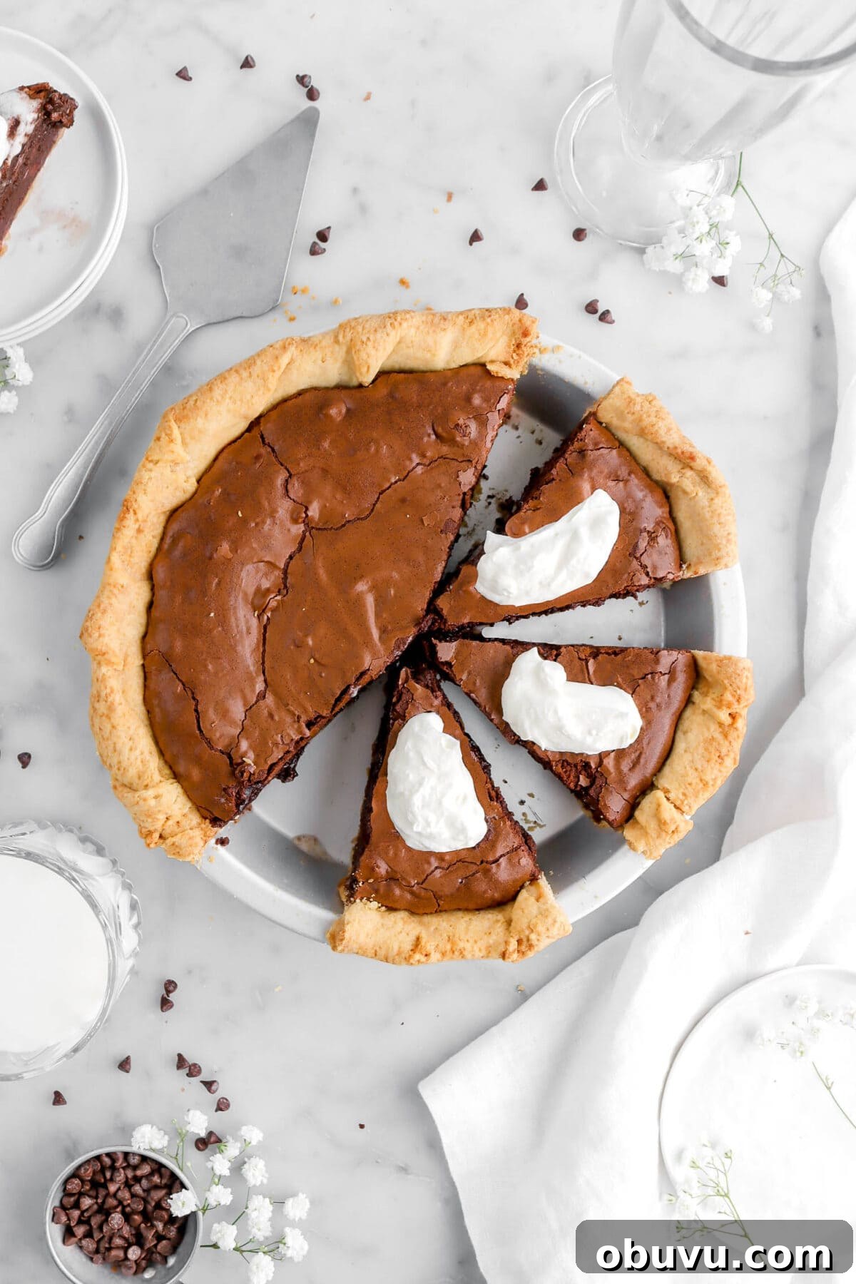Fudgy Chocolate Chip Brownie Pie Recipe for Ooey-Gooey Dessert 6 close up overhead shot of brownie pie with three slices cut into pie with whipped cream on each piece, chocolate chips around and white flowers.
