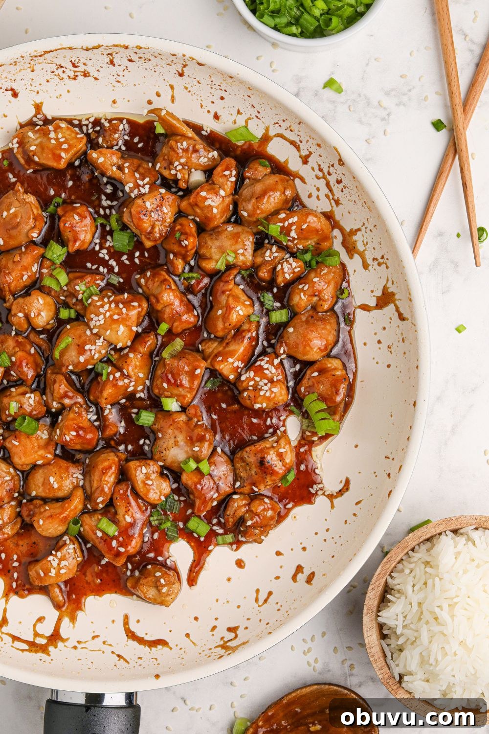30-Minute Bourbon Chicken Skillet Recipe That Beats Takeout 18 a white skillet full of saucy bourbon chicken on a table with chopsticks to the side and a bowl of rice to the side of the pan