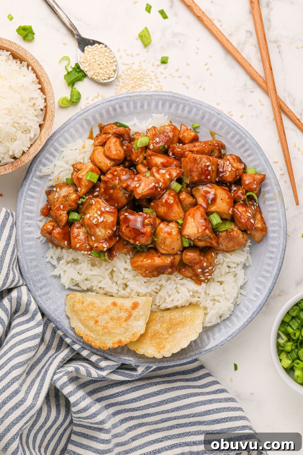30-Minute Bourbon Chicken Skillet Recipe That Beats Takeout 21 looking down on table with a grey plate with bourbon chicken, rice, and pieorgies to side and a bowl of rice and green onions to side on the table