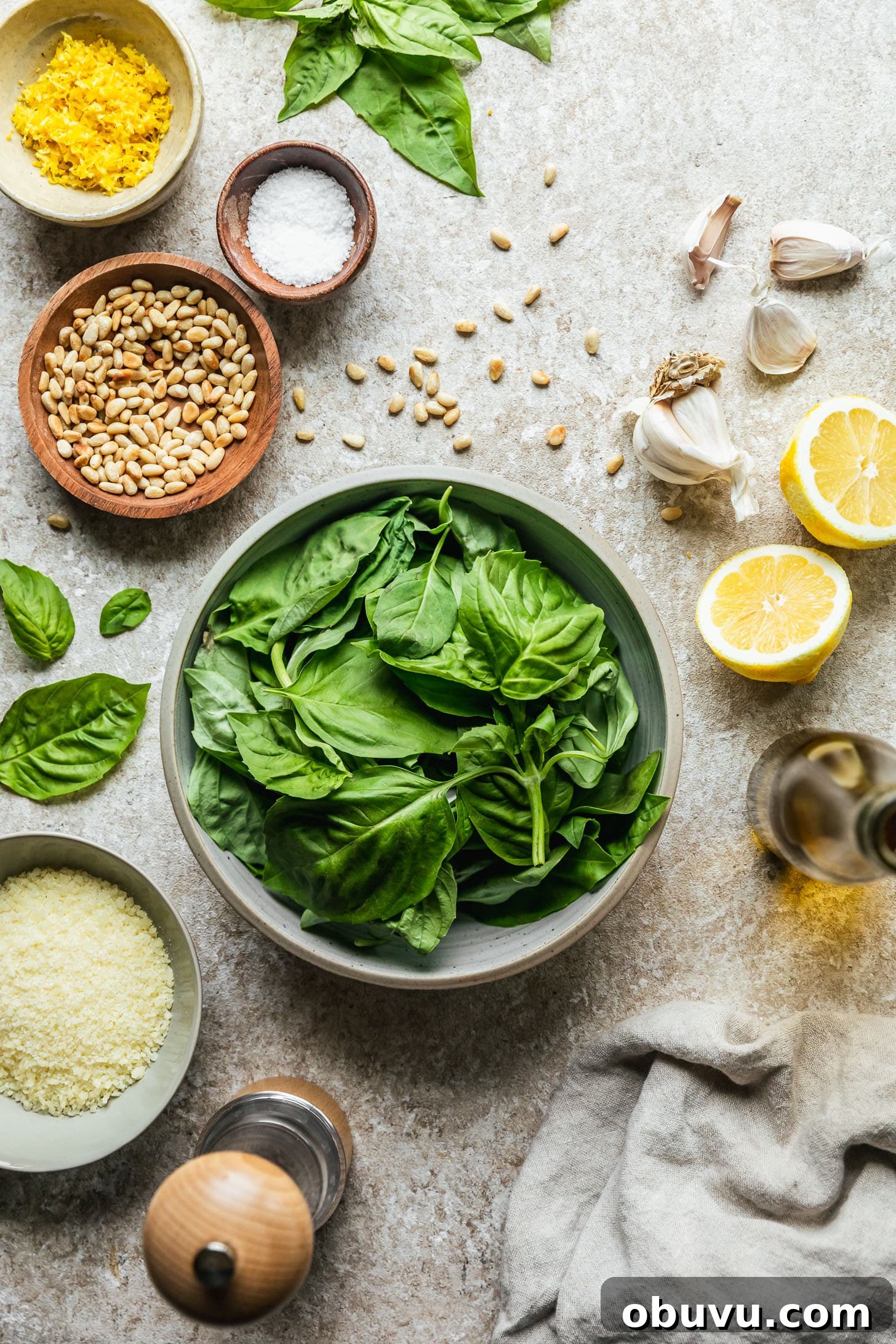 A grey bowl of basil on a beige counter next to halved lemons, a jar of olive oil, a beige linen, a grey bowl of Parmesan, a white bowl of lemon zest, and a wood bowl of pine nuts.