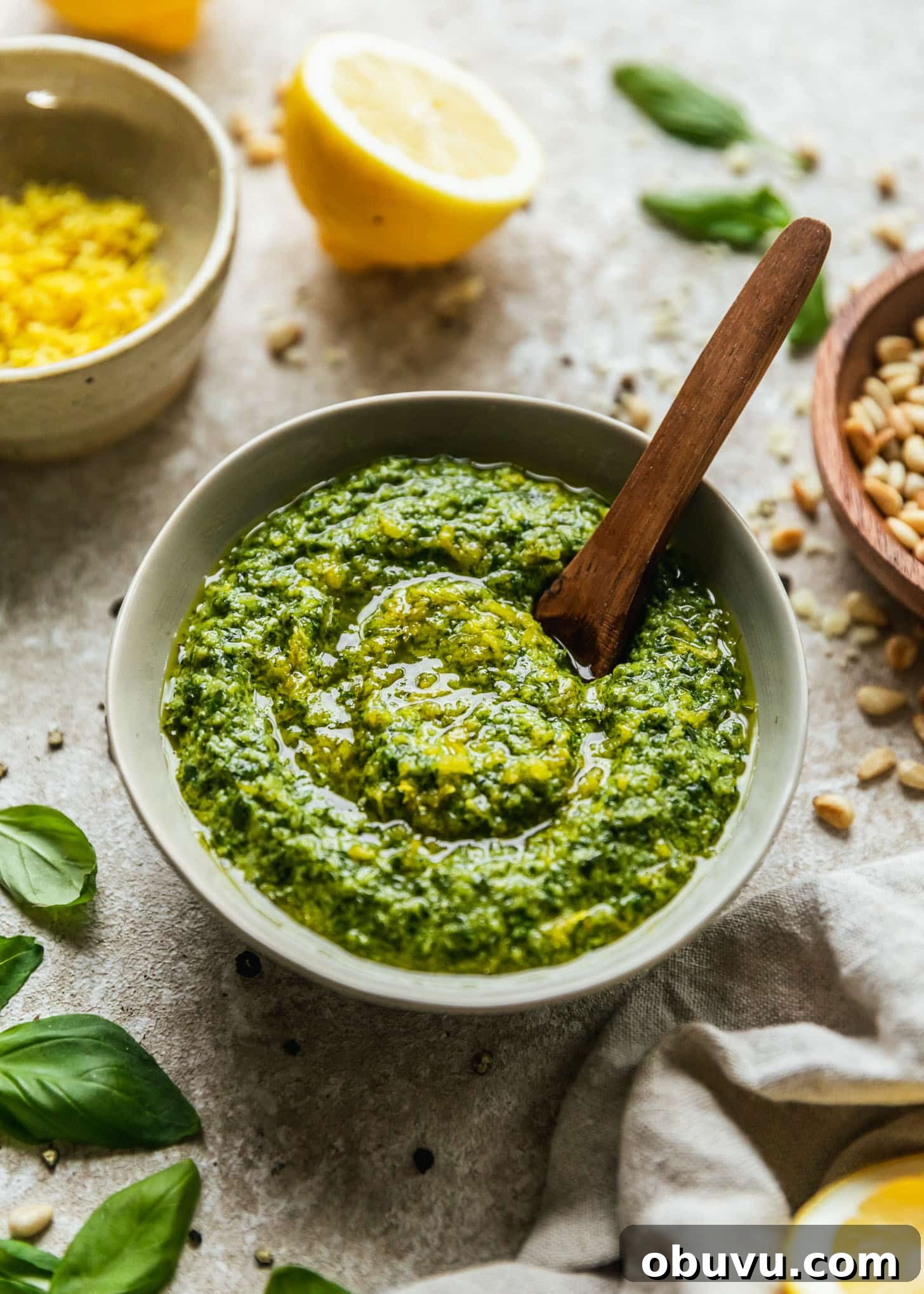 A grey bowl of lemon basil pesto on a beige counter next to halved lemons, a wood bowl of pine nuts, a white bowl of lemon zest, and basil leaves.