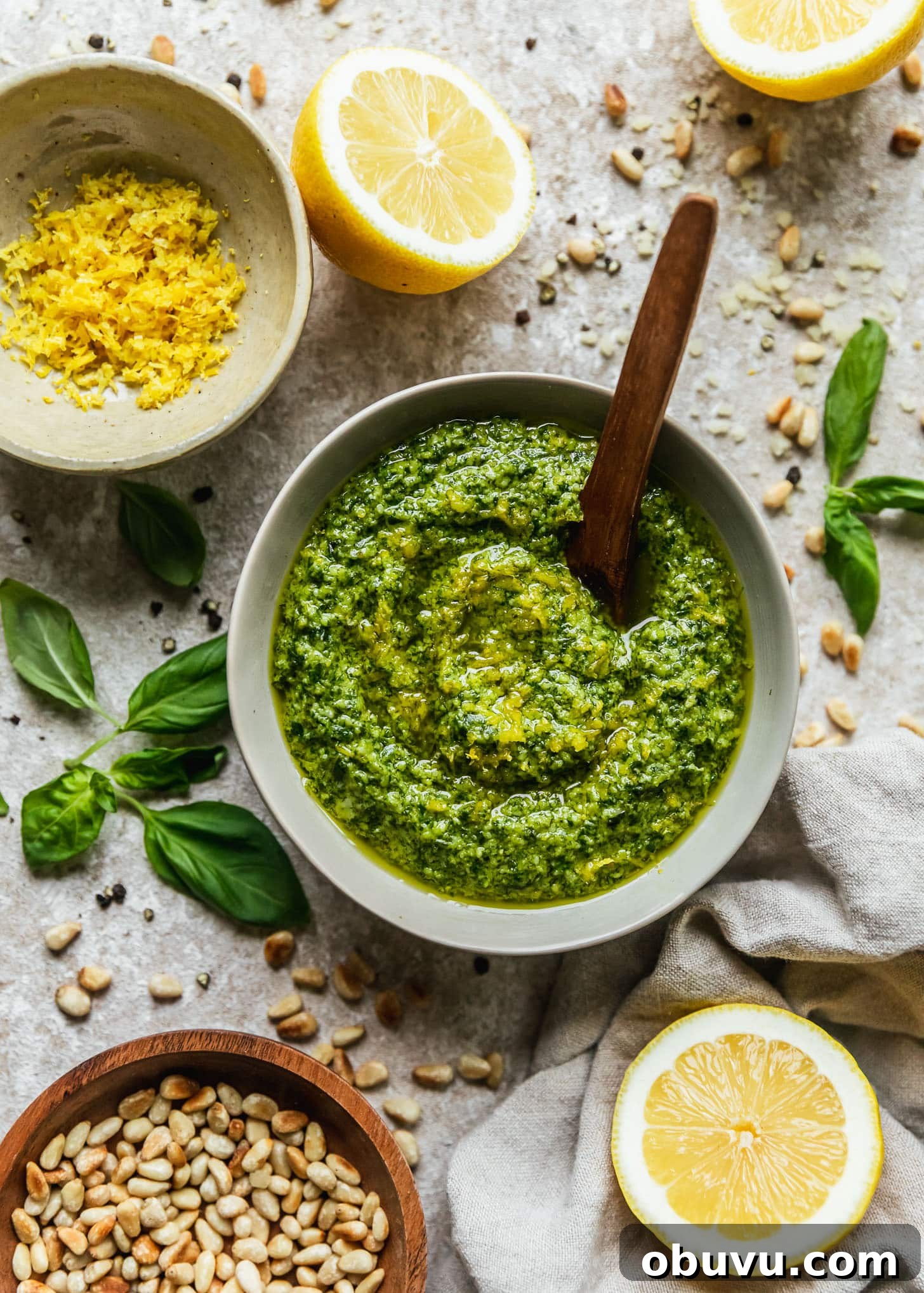A grey bowl of lemon pesto with a wooden spoon on a beige counter next to lemon halves, basil leaves, a white bowl of lemon zest, a wood bowl of pine nuts, and a beige linen.
