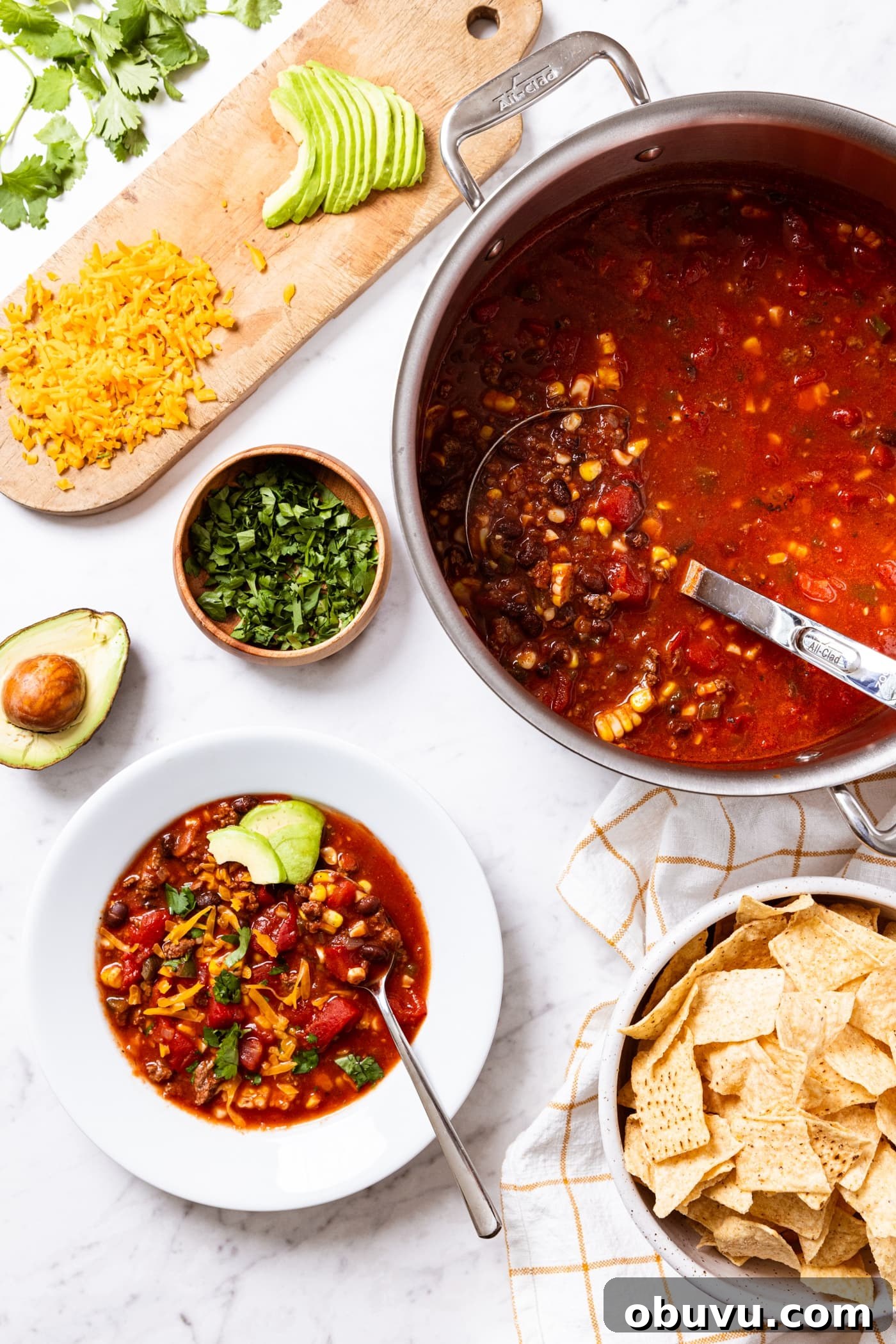 Quick Homemade Taco Soup Recipe for Cozy Weeknight Dinners 3 White marble surface with all components of an easy taco soup including hot red colored taco soup and white bowl filled with serving of soup fresh from the stockpot with toppings all spread around to put on top of the soup.