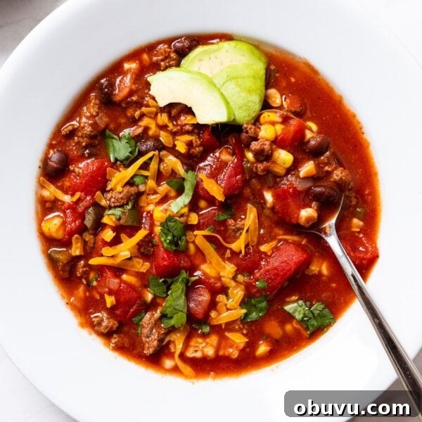 Quick Homemade Taco Soup Recipe for Cozy Weeknight Dinners 6 White serving bowl filled with single serving of taco soup topped with cheese and slices of green avocado with spoon resting in bowl all on white marble surface.