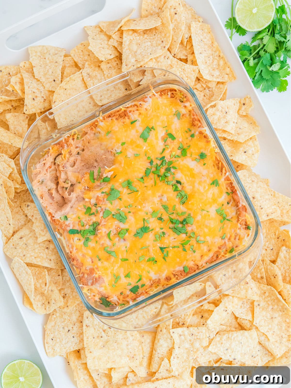 Cheesy Baked Refried Bean Dip for Parties and Game Day 2 Square glass dish of cheesy bean dip garnished with herbs, surrounded by tortilla chips on a tray. Lime and cilantro are placed in the corner.