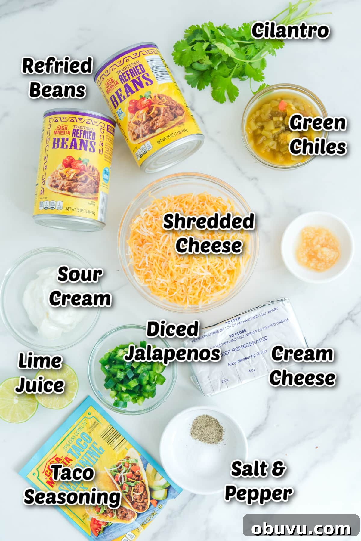 Cheesy Baked Refried Bean Dip for Parties and Game Day 3 Ingredients on a table: refried beans, cilantro, green chiles, shredded cheese, sour cream, lime juice, diced jalapenos, cream cheese, taco seasoning, salt, and pepper.