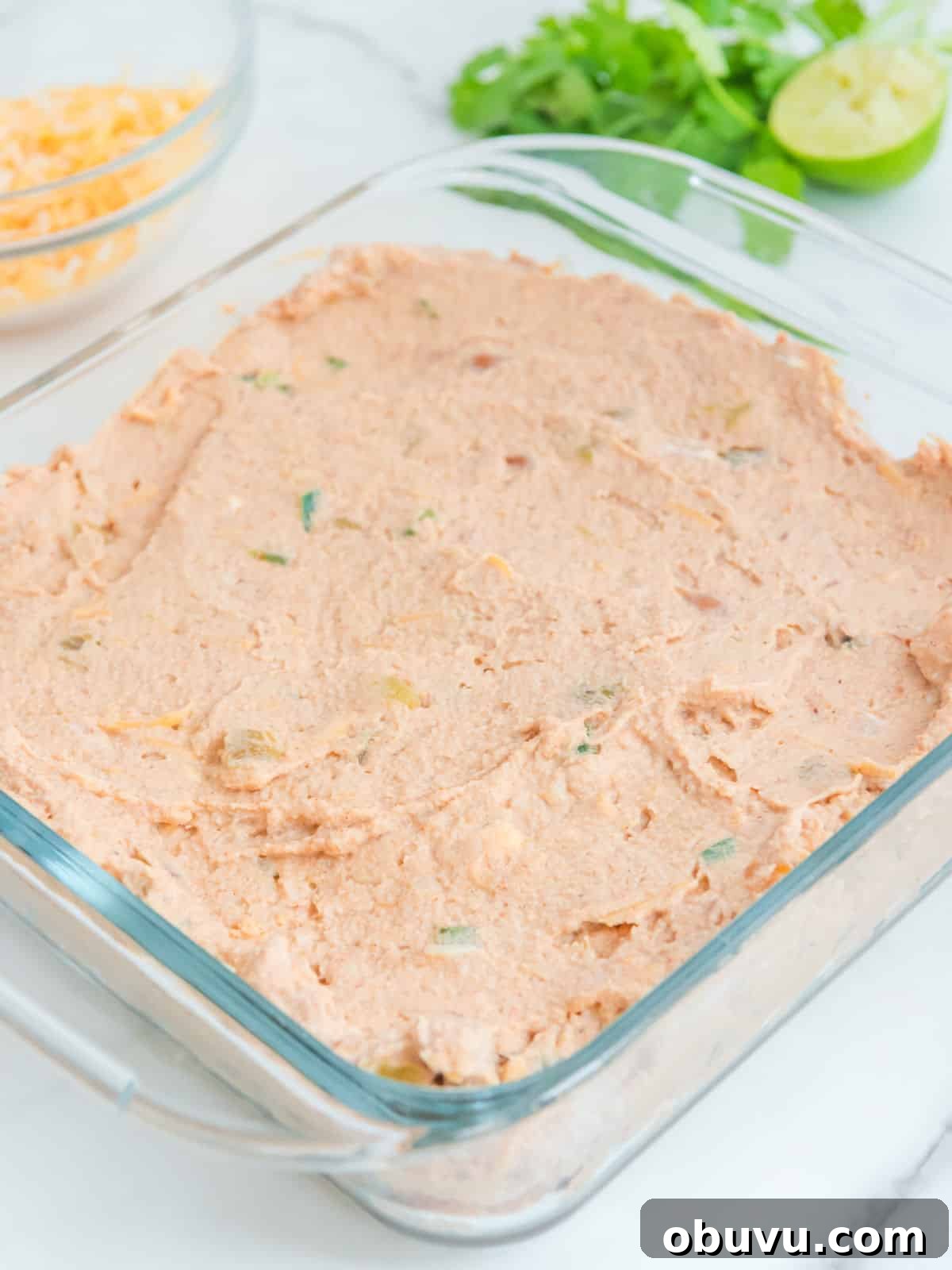 Cheesy Baked Refried Bean Dip for Parties and Game Day 5 A glass square dish filled with a light orange bean dip with herbs, on a marble surface. A bowl of cheese, chopped cilantro, and a sliced lime are in the background.