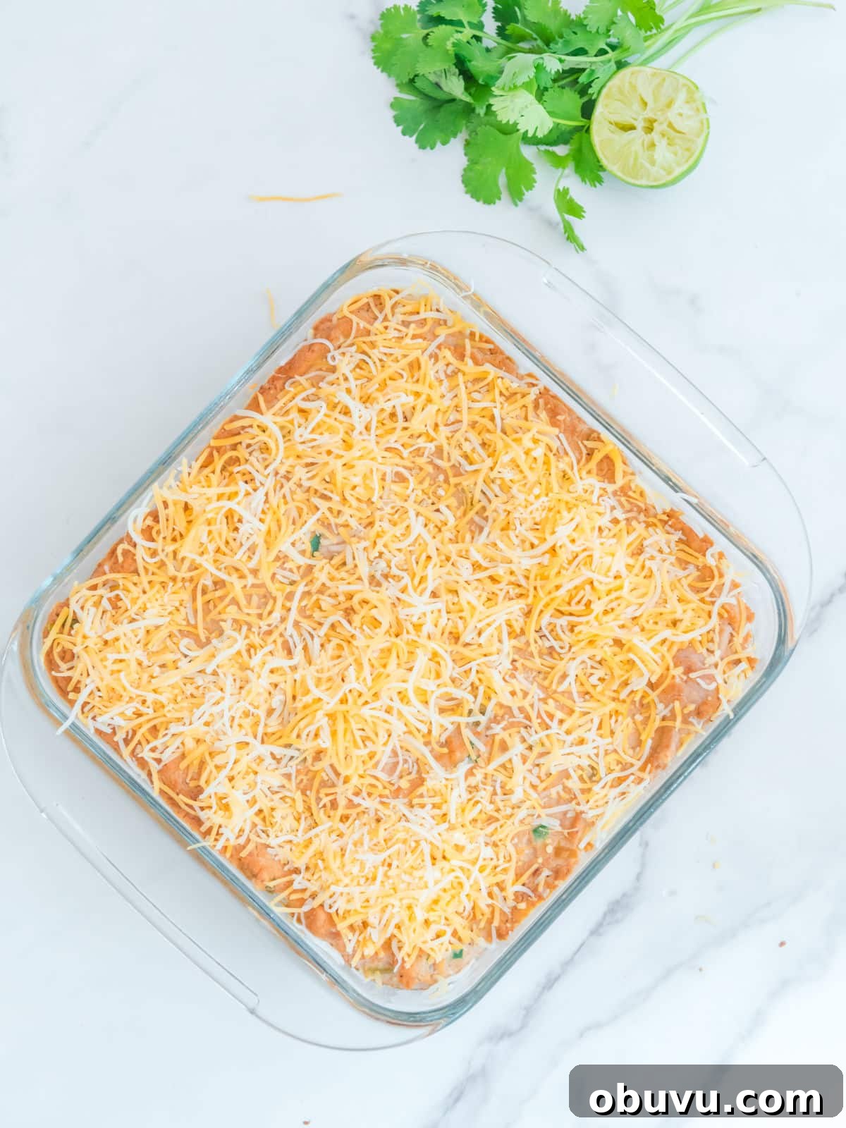 Cheesy Baked Refried Bean Dip for Parties and Game Day 6 Glass dish of shredded cheese on top of a baked dish, garnished with cilantro and lime on the side.