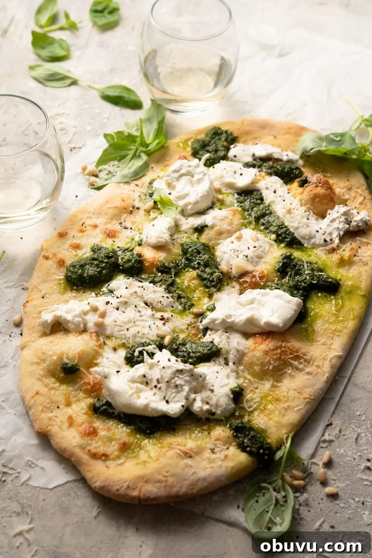 Cheesy Basil Pizza with Burrata and Pesto A basil pizza with burrata and pesto on a wooden surface with glasses of white wine in the background