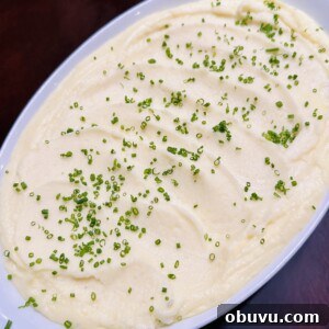 A white oval dish filled with creamy mashed potatoes, topped with finely chopped chives and set on a dark wooden table.