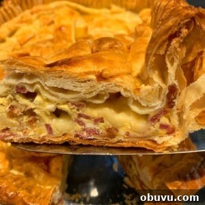 A close-up of a slice of pie being served, showcasing layers of flaky golden-brown pastry filled with creamy cheese and pieces of ham. The rest of the pie is in the background.