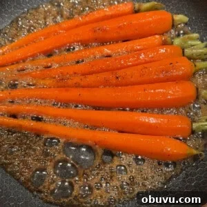 Bright orange carrots being sautéed in a frying pan with bubbling brown sauce. The carrots are evenly spaced, and the sauce appears to be well-seasoned with black pepper.