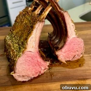 A perfectly cooked rack of lamb cut into two sections, displaying a pink, juicy interior and seasoned crust, resting on a wooden cutting board in a kitchen setting.