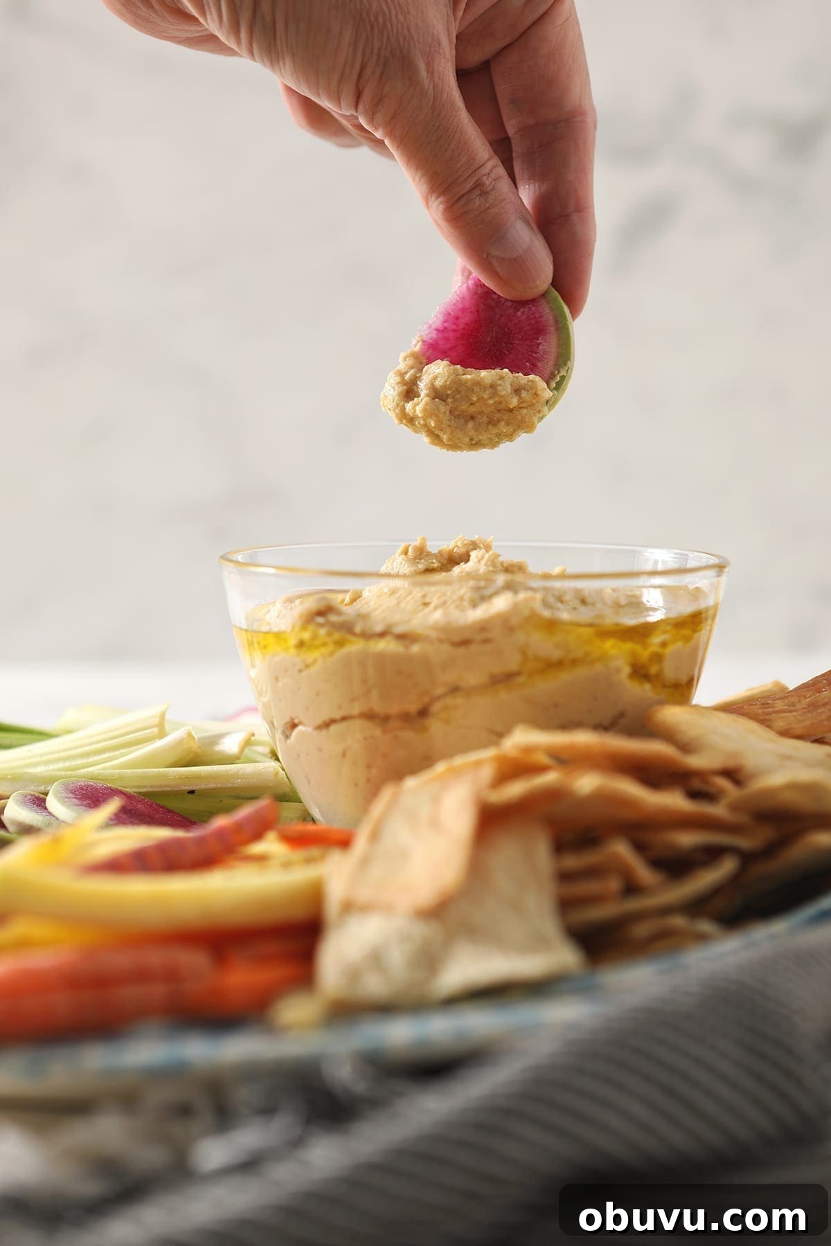 Winston’s Roasted Garlic Hummus Recipe 7 A woman dips a watermelon radish into a clear glass bowl of homemade garlic hummus
