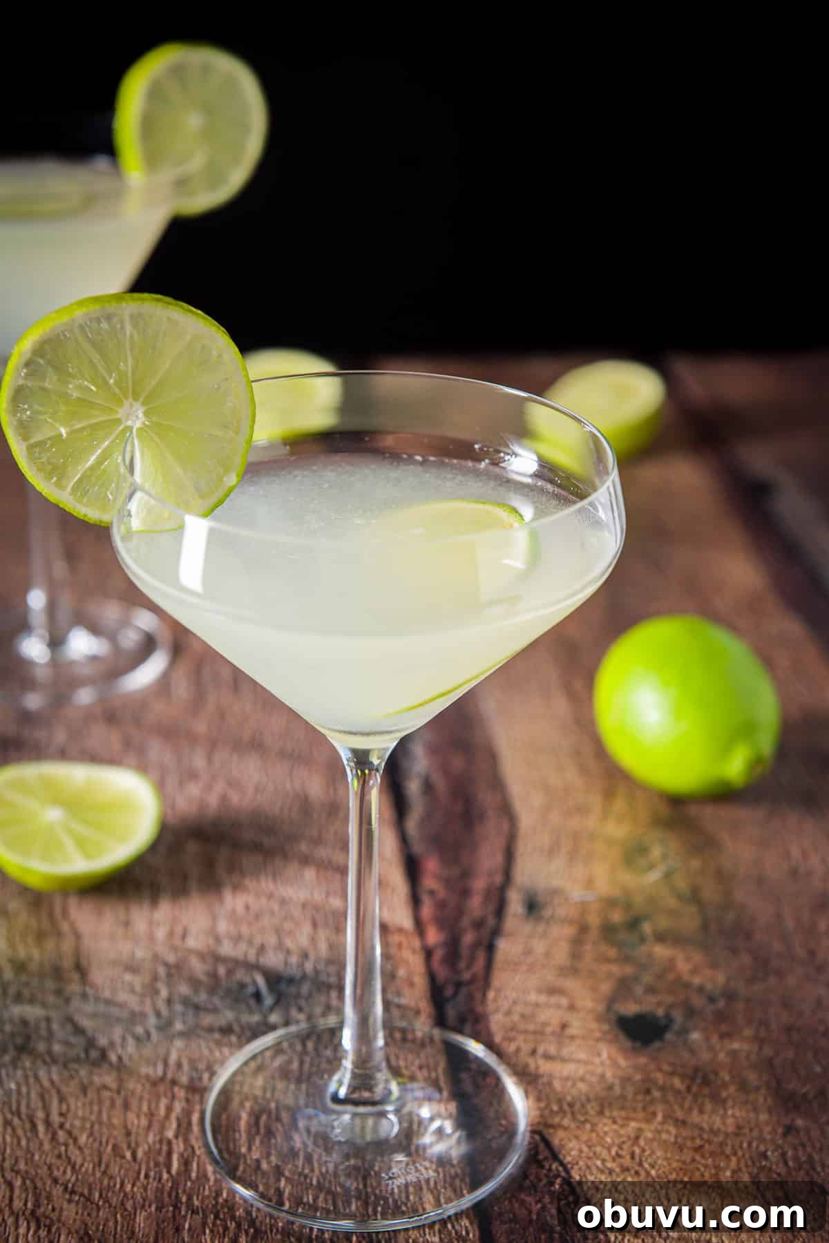 Classic Kamikaze Cocktail Recipe and Serving Tips 6 A curved martini glass with lime liquid garnished with lime wheels. There are also limes on the table and another glass in the background