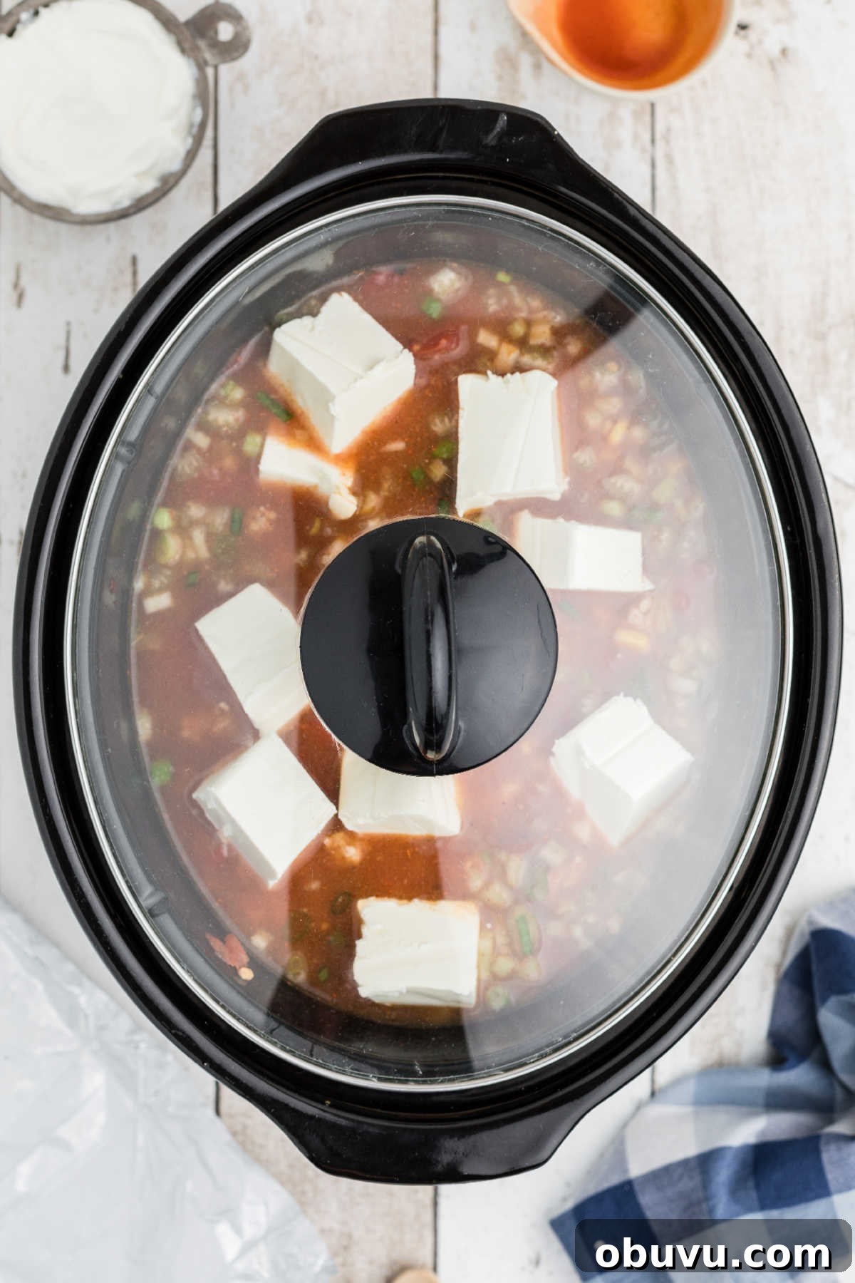 A slow cooker with a lid on, covering some buffalo chicken chili, with cream cheese melting into it.
