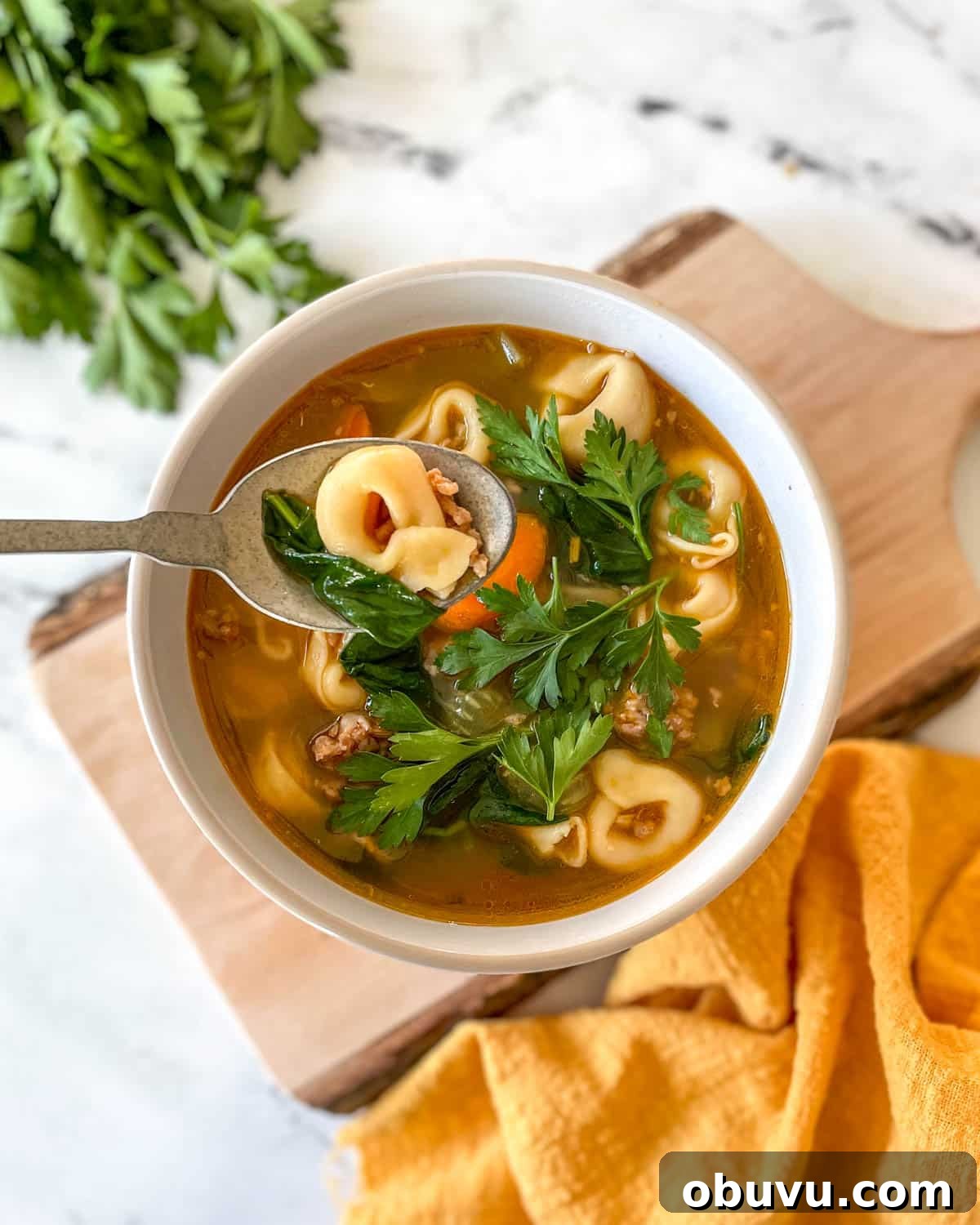 A bowl of tortellini soup sits on a rustic wooden cutting board with a yellow linen and a bunch of parsley.