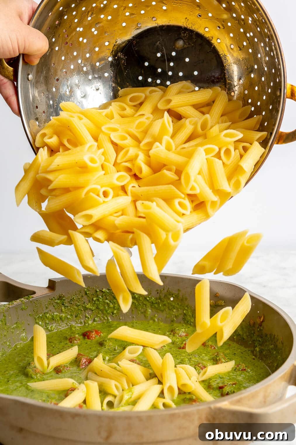 Salmon Pesto Pasta Recipe: Creamy 20-Minute Meal 9 A person pouring penne pasta from a colander into a pan of pesto sauce.