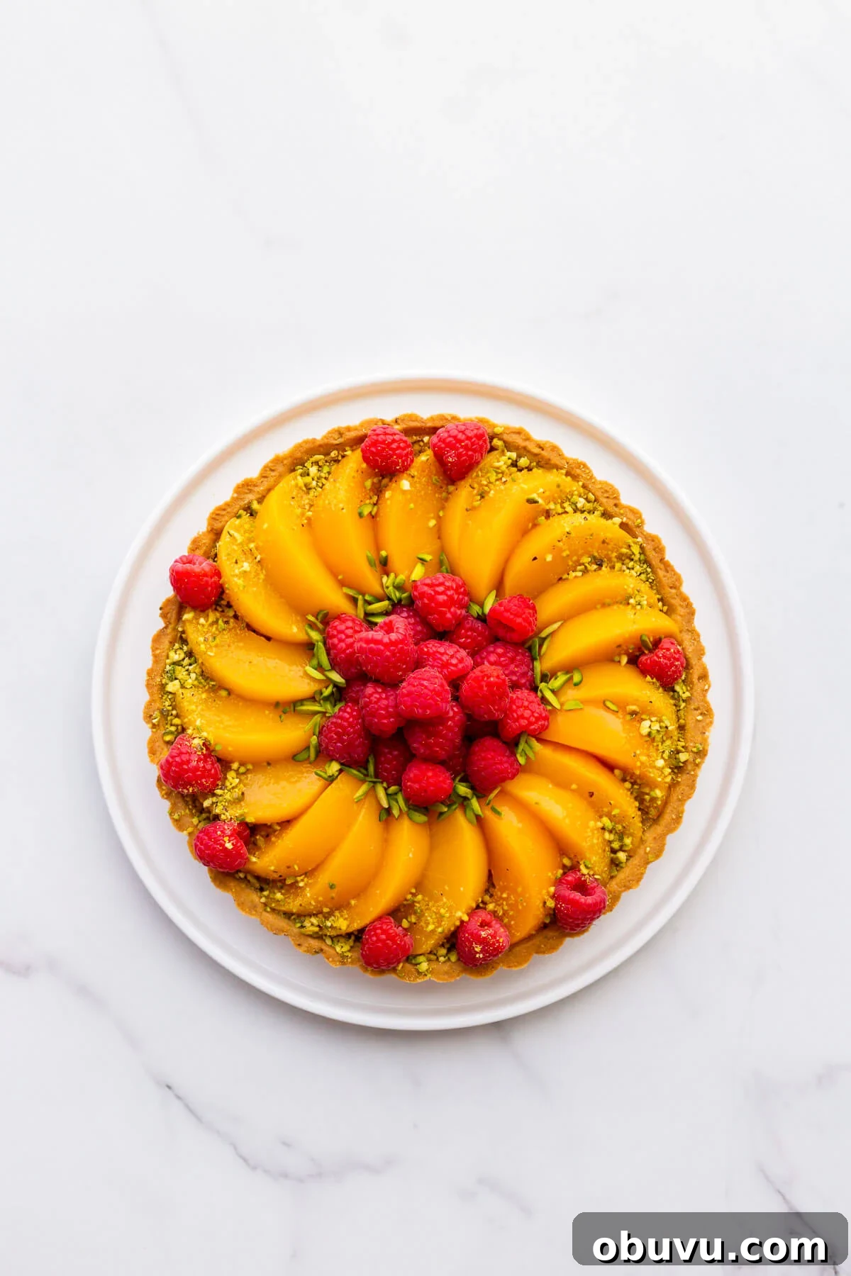 Pistachio tart topped with sliced peaches and fresh raspberries, and decorated with slivered and ground green pistachios.