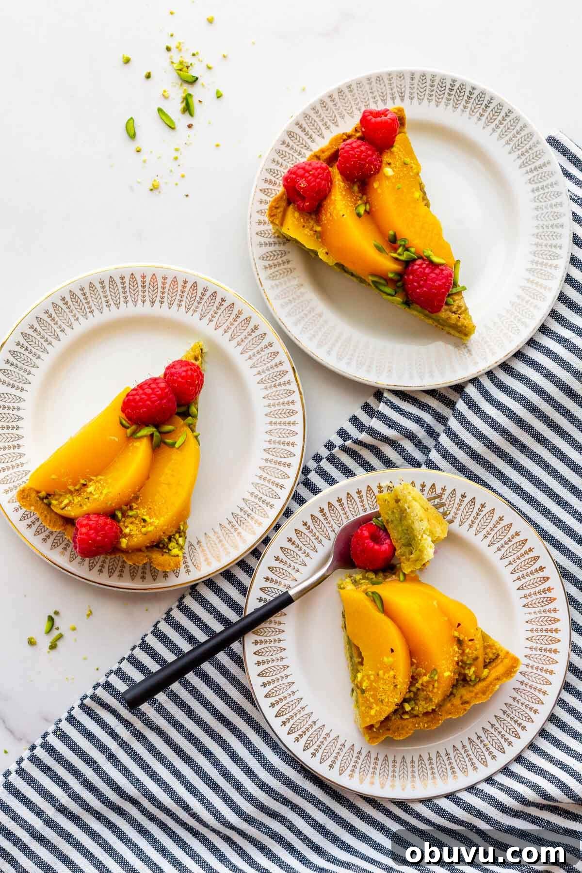 Three slices of pistachio tart with raspberries and peaches on plates.