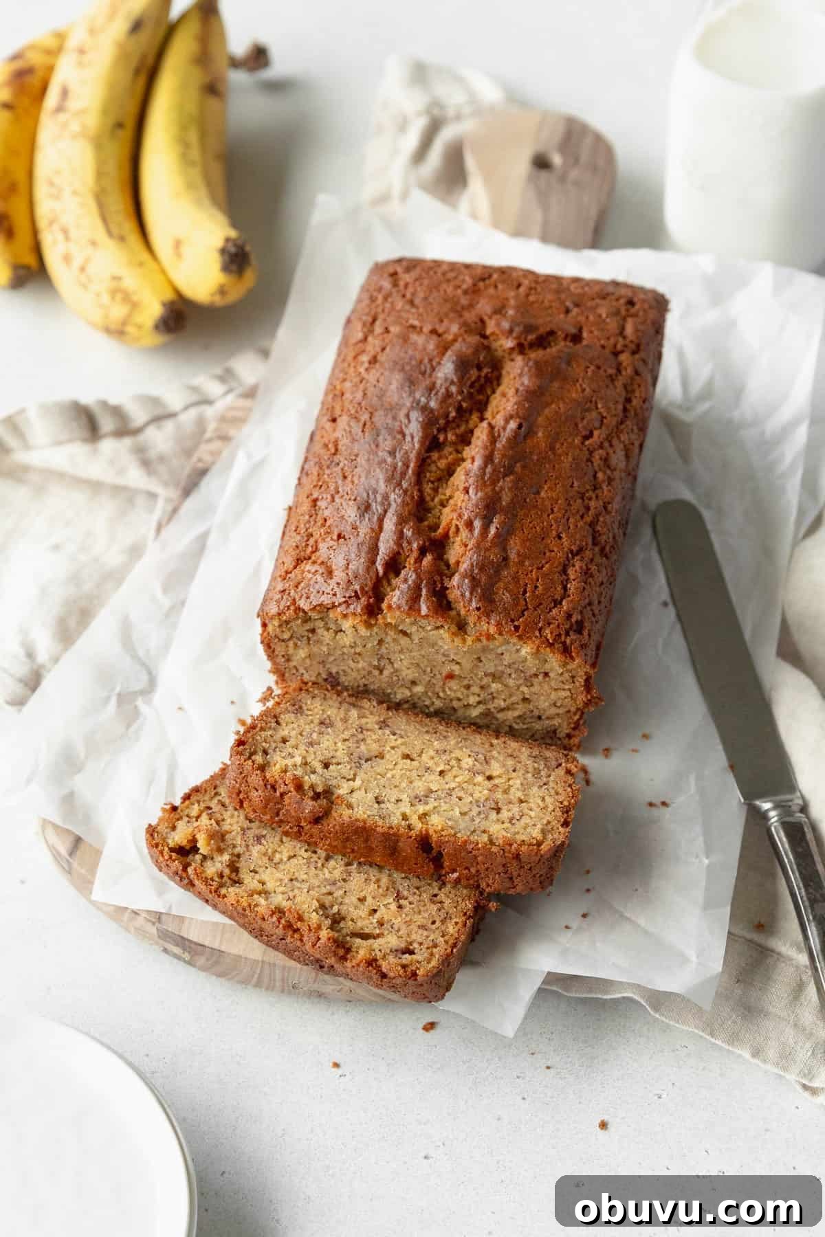 Moist Banana Bread Without Gluten — Simple Recipe for Tasty Loaf 2 A sliced loaf of gluten free banana bread on sheets of parchment paper with a bunch of banana in the background.