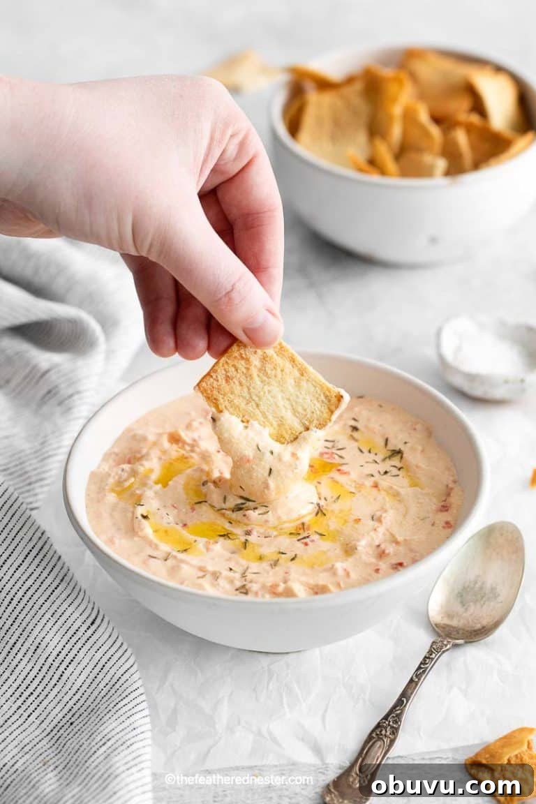 a hand holding a pita chips dipping it to the Mediterranean-inspired roasted red pepper feta dip on a bowl with a spoon beside it