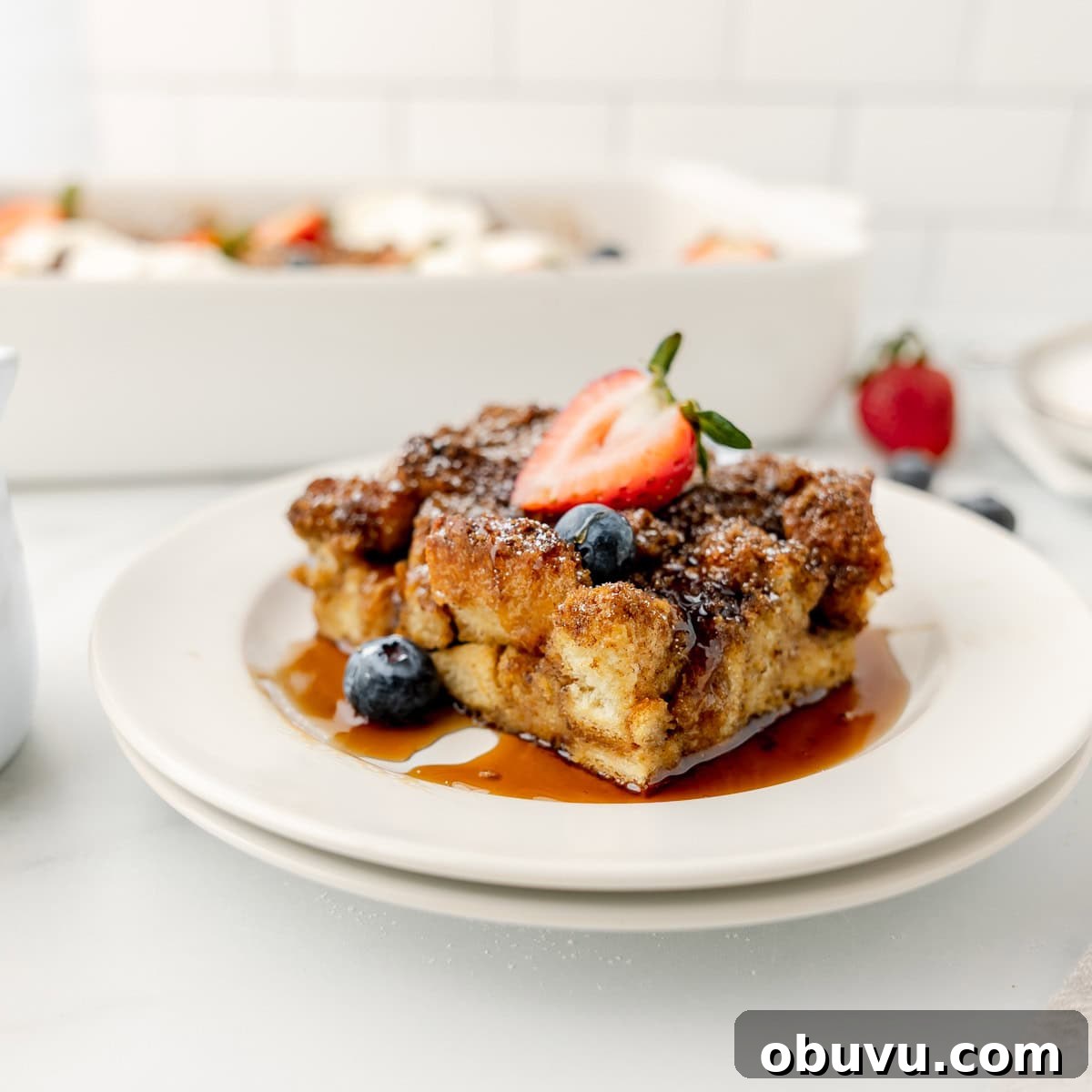Overnight Brioche French Toast Casserole Recipe 2 A slice of baked French toast topped with syrup, a strawberry, and blueberries on a white plate.
